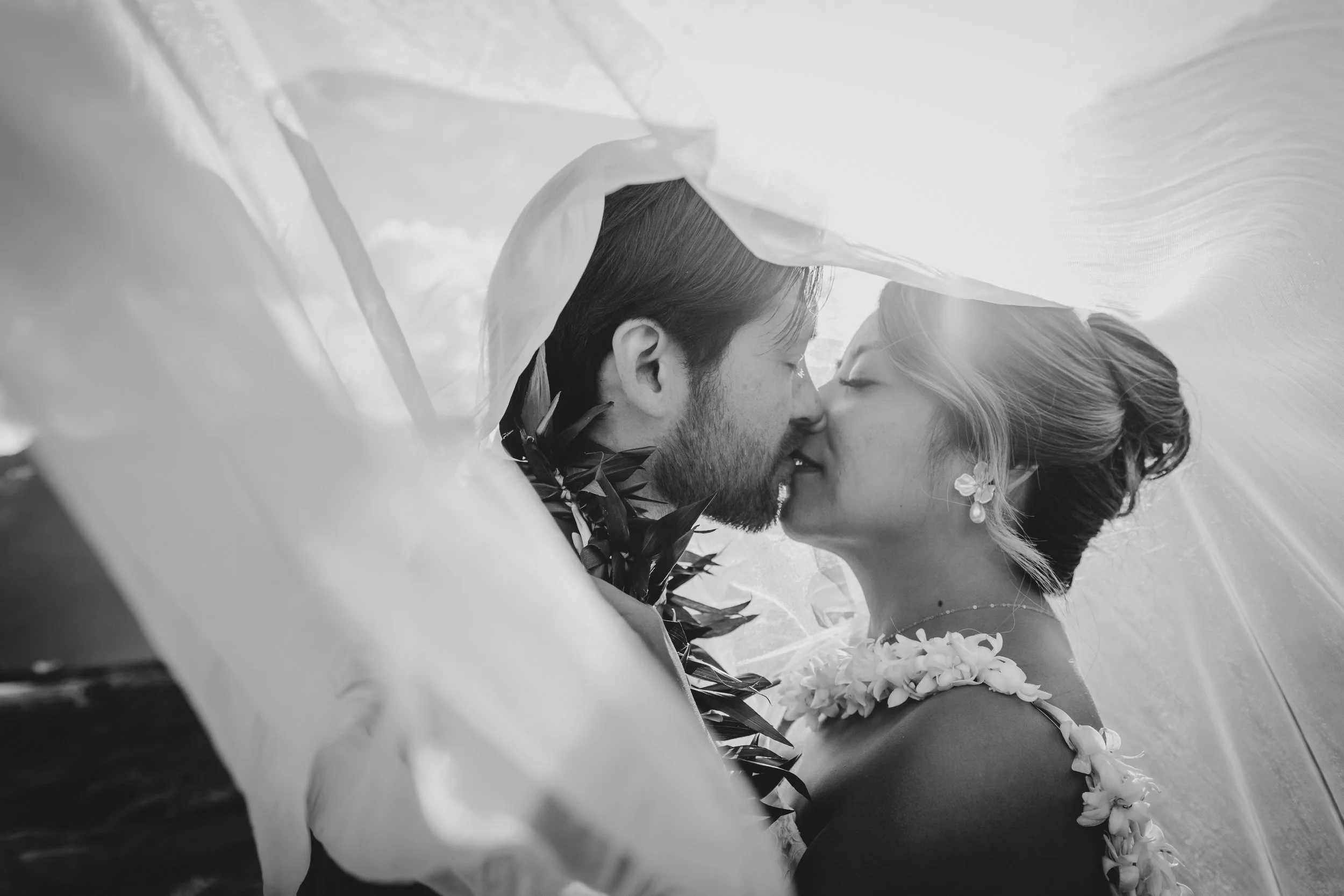A black and white photograph of a couple kissing under a large, flowing fabric or veil, with a floral lei around the woman's neck, suggesting a wedding or romantic moment.