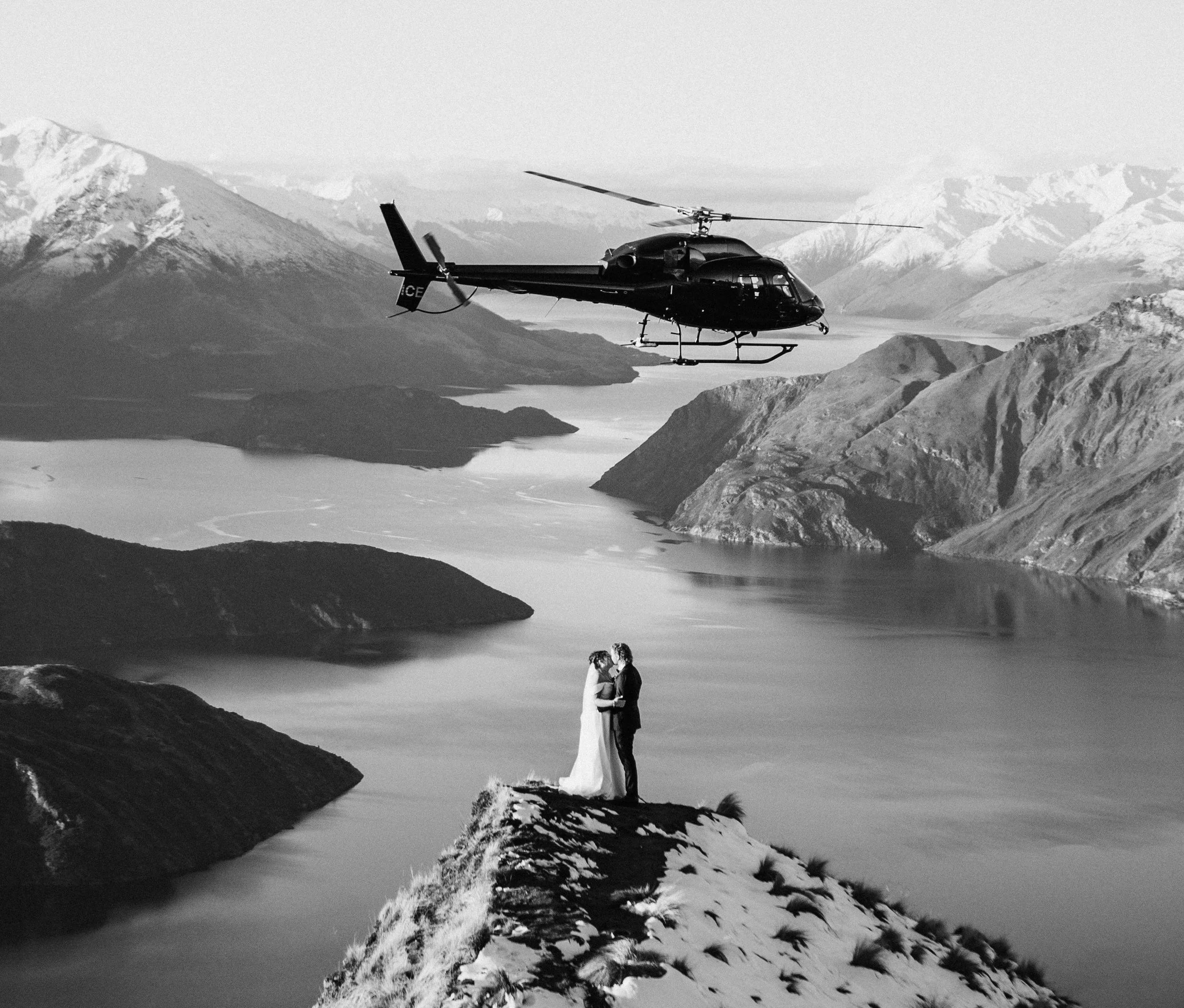A couple in wedding attire standing on a rocky ledge overlooking a body of water in a mountain landscape, with a helicopter flying nearby.