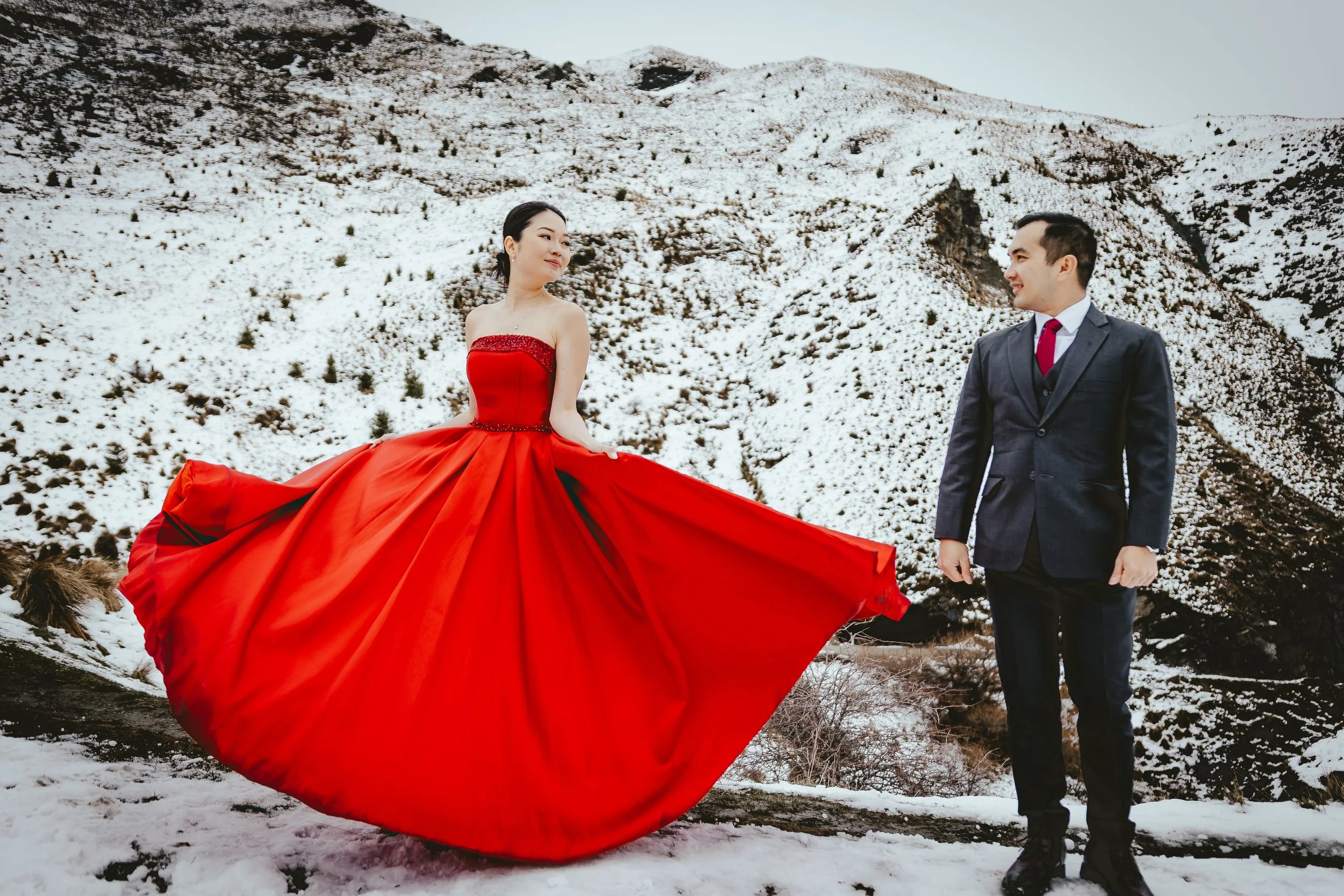 A woman in a red strapless gown with a flowing skirt stands in the snow with a man in a gray suit, white shirt, and red tie, with snow-covered mountains in the background.