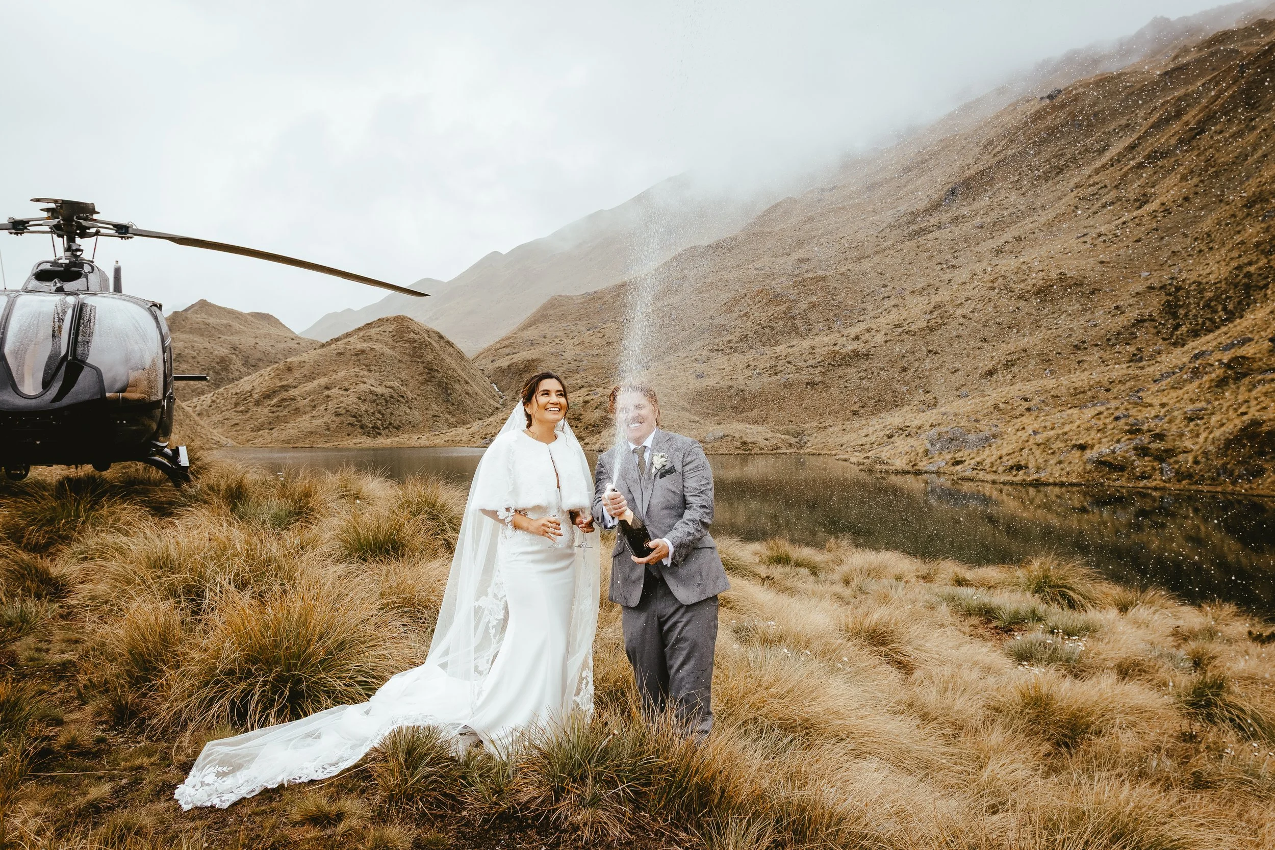 A bride and groom celebrating outdoors near a lake with mountains in the background, a helicopter with a camera is nearby, and the groom is opening a bottle of champagne.
