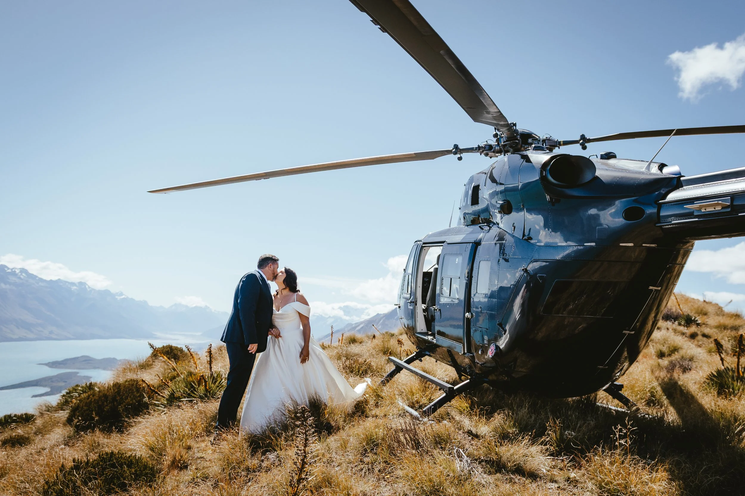 A bride and groom stand on a grassy hillside in front of a black helicopter, with a lake and mountains in the background, during daytime.