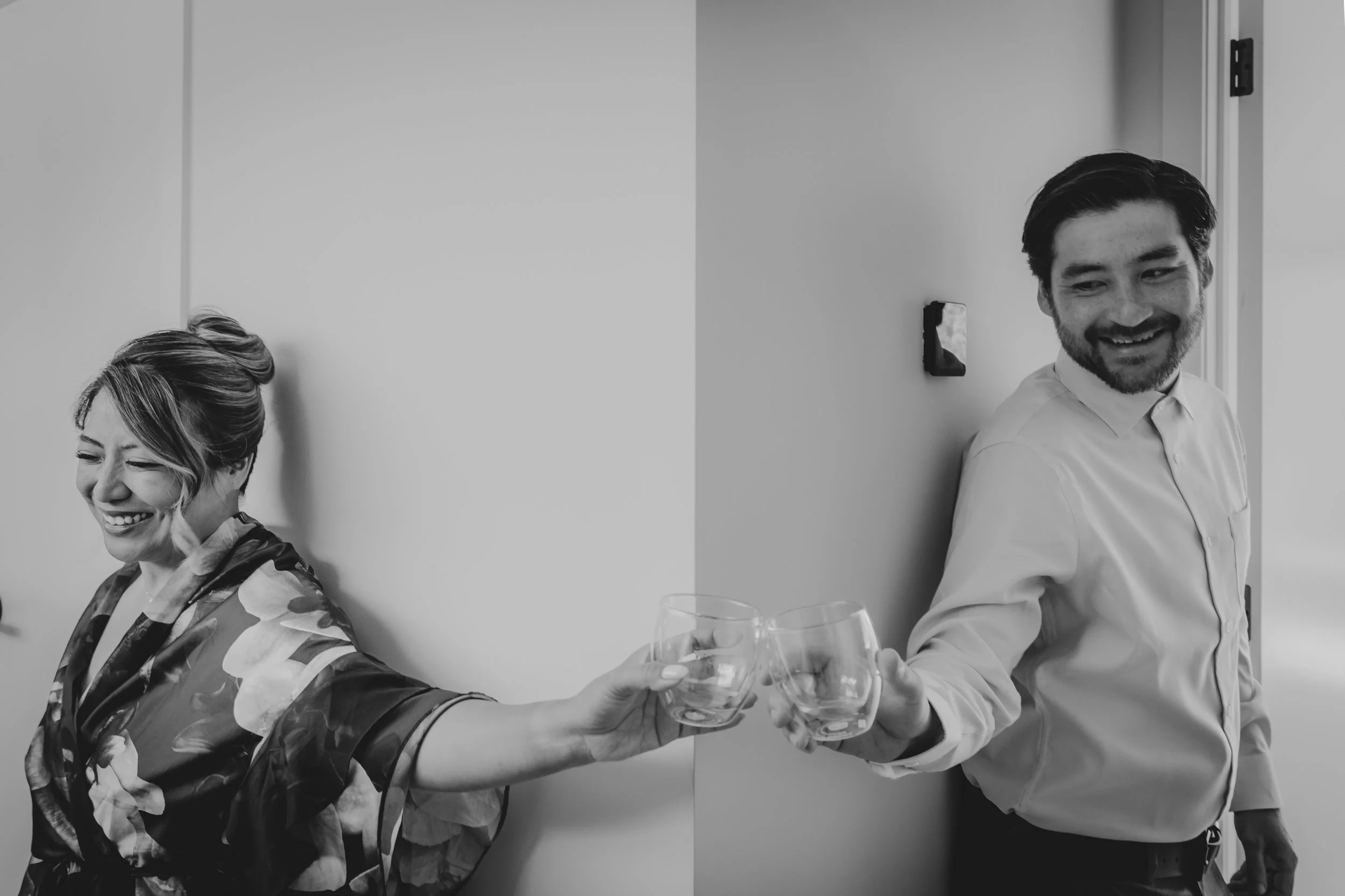 A woman and a man are reaching across a white wall to clink glasses of wine, smiling.