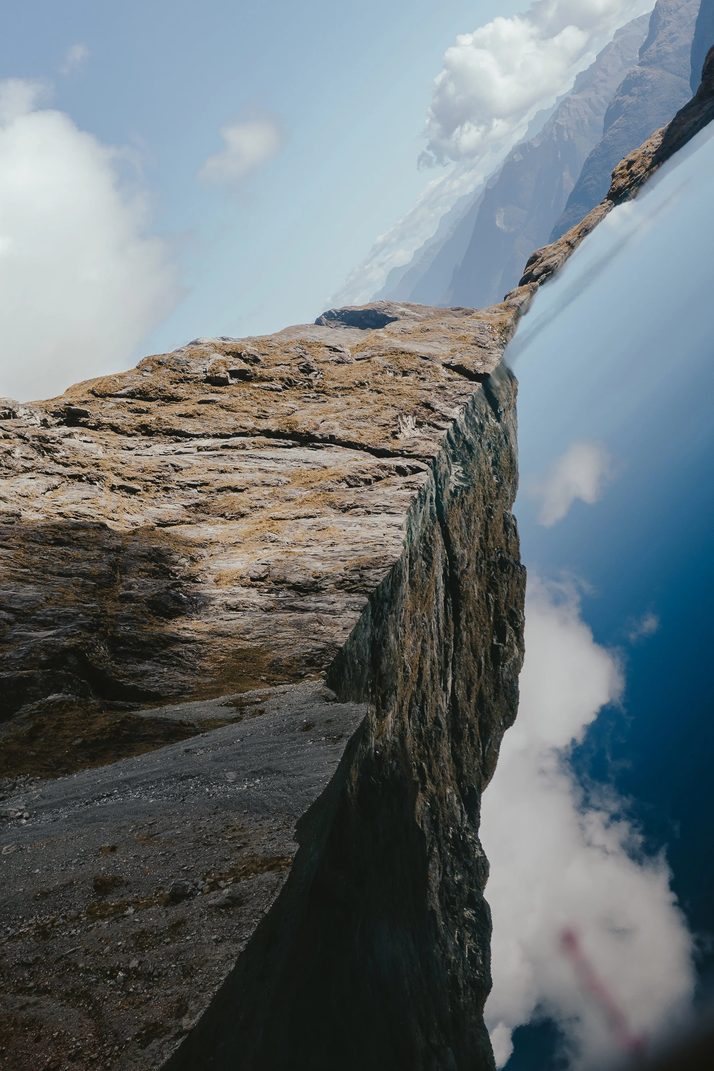 A rugged mountain path carved along a steep cliffside, with a dramatic sky filled with clouds and distant mountains in the background.