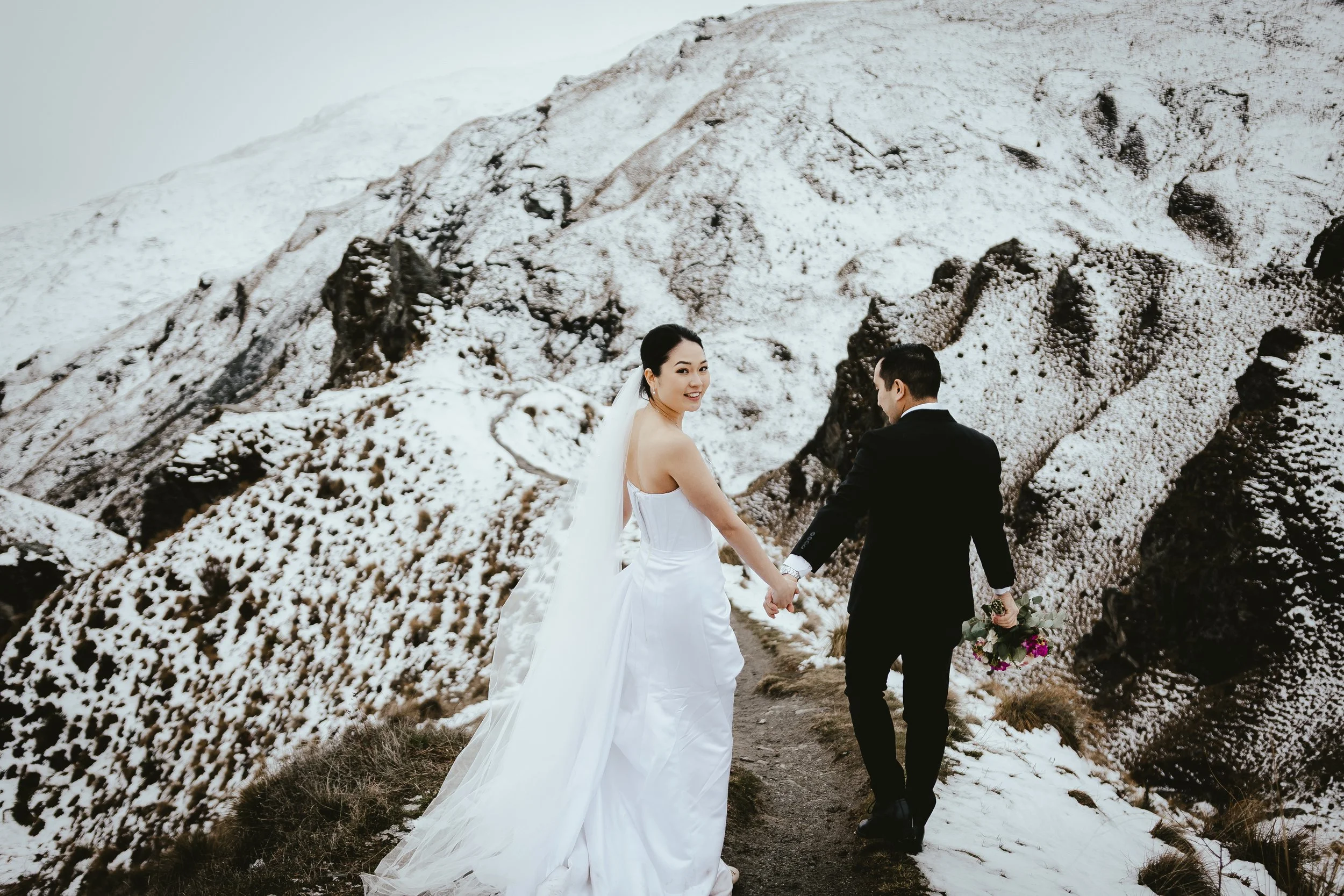 A bride and groom holding hands and walking on a snowy mountain trail during their wedding, with the bride smiling at the camera and the groom holding a bouquet of flowers.
