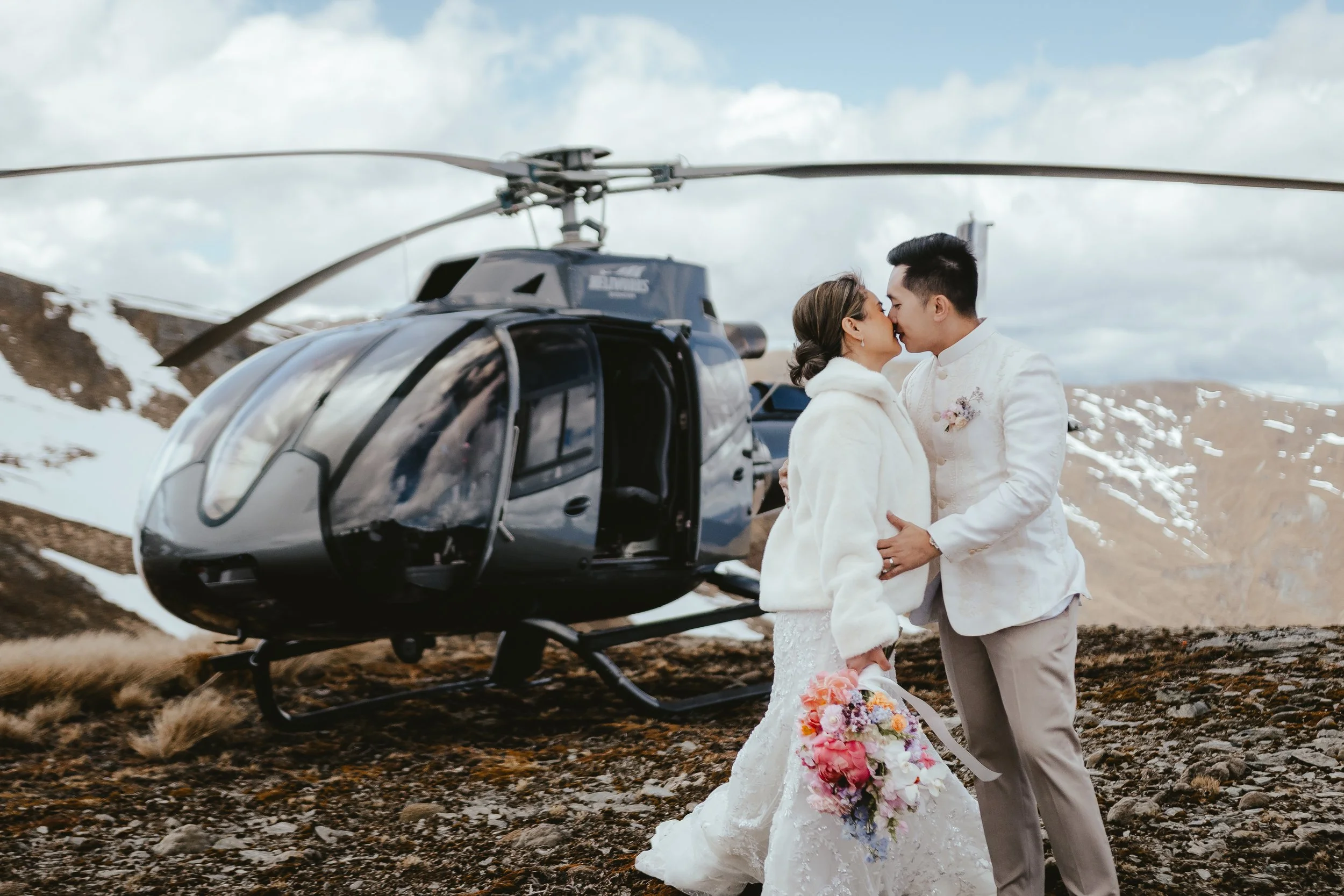 A bride and groom sharing a kiss outdoors on a mountain landscape with patches of snow, standing next to a black helicopter.