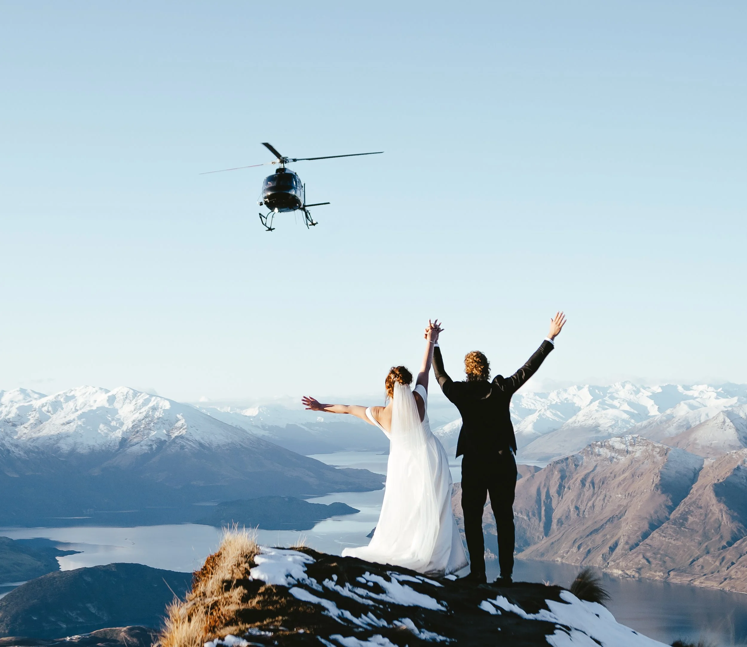 A bride and groom standing on a mountain overlooking snowy peaks and a lake, with a helicopter flying nearby against a clear sky.