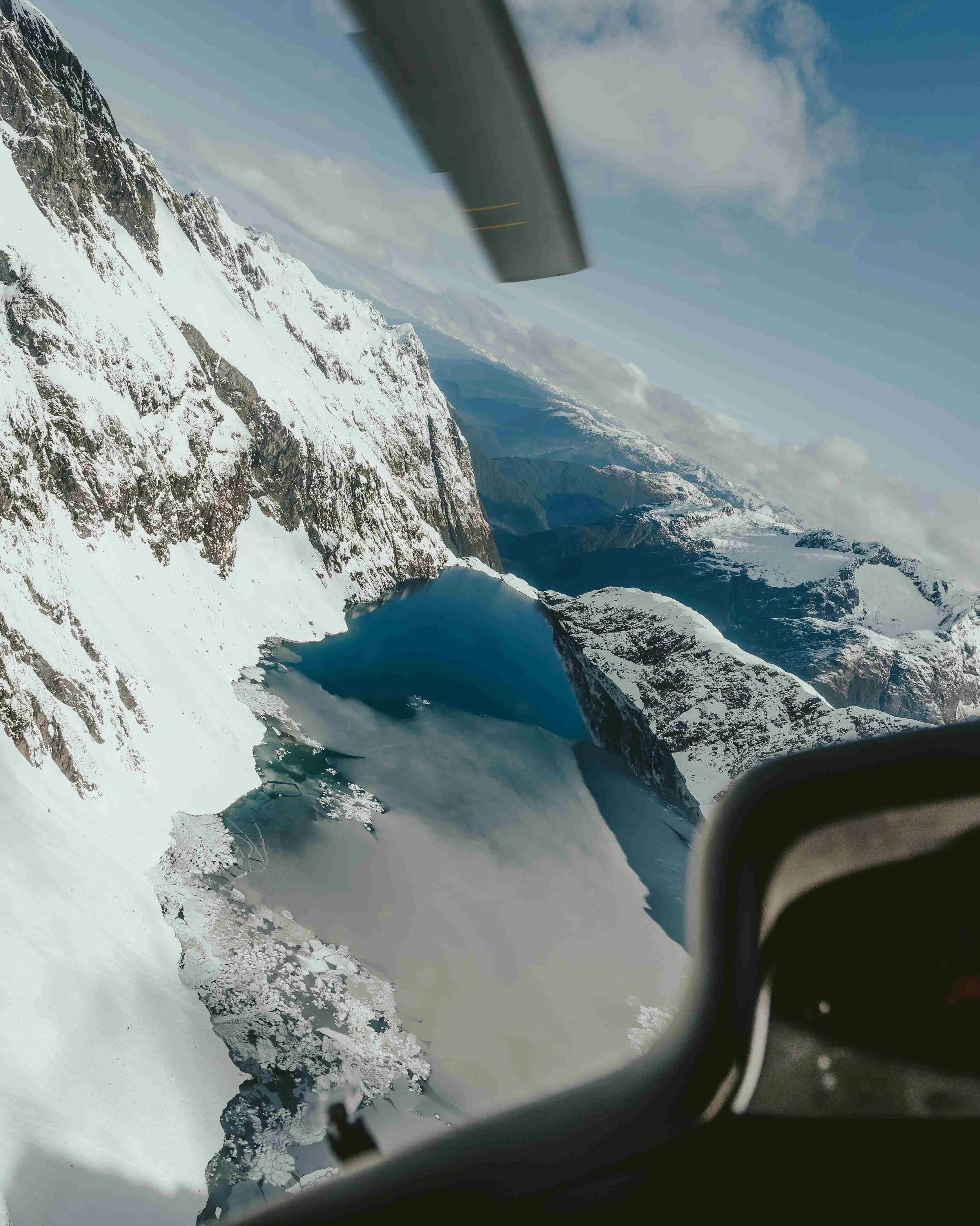 Snow-covered mountains seen from an airplane window with part of the aircraft's wing and engine visible in the foreground.