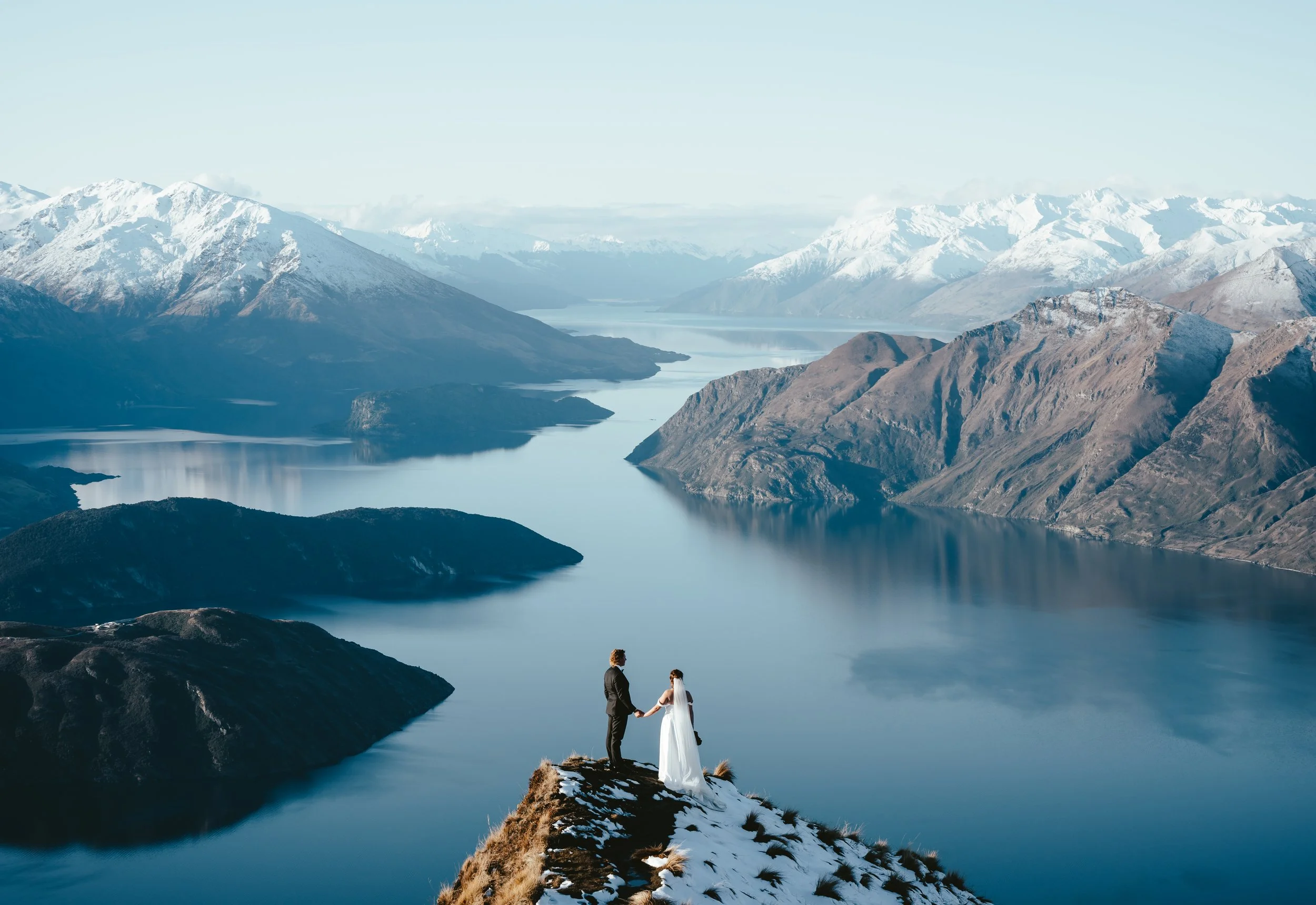 A couple dressed in wedding attire holding hands on a rocky outcrop overlooking a lake and snow-capped mountains.