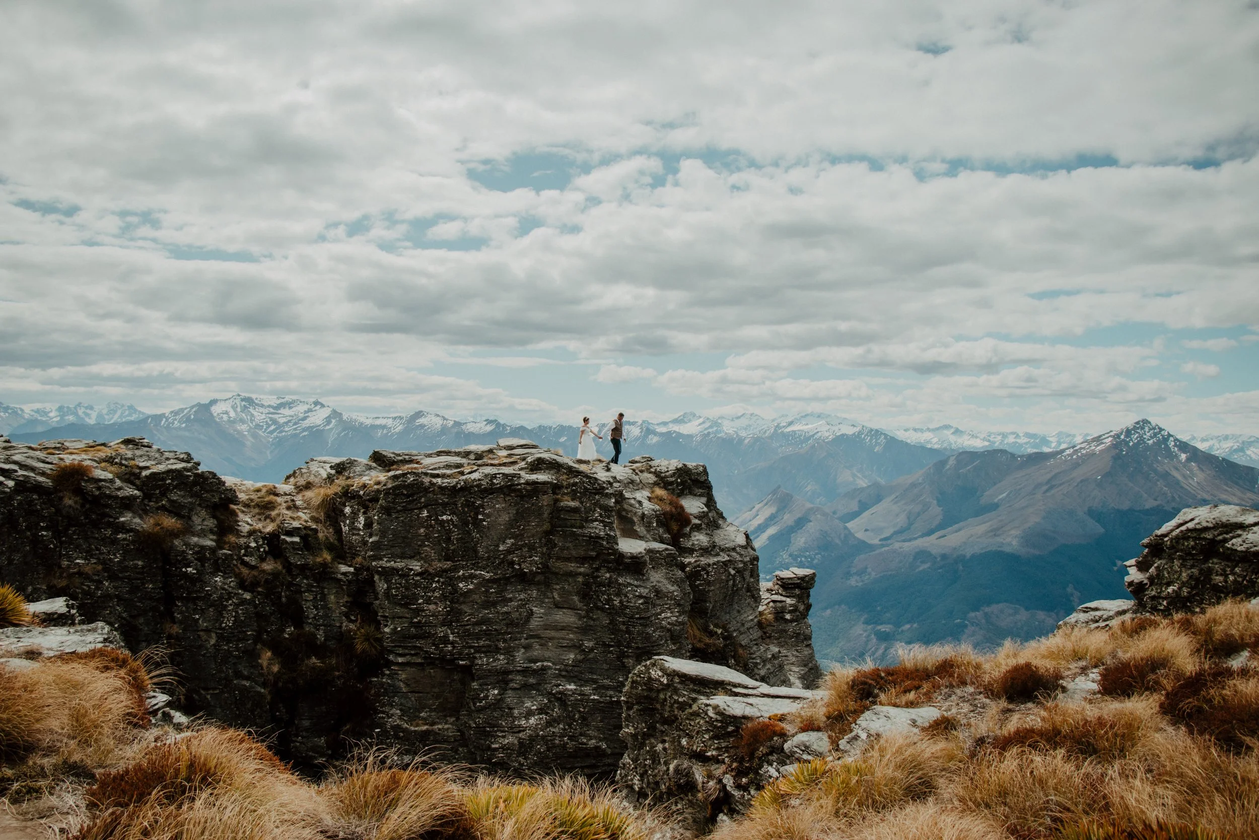 A couple holding hands standing on rocky terrain with mountainous landscape and snow-capped peaks in the background under a cloudy sky.