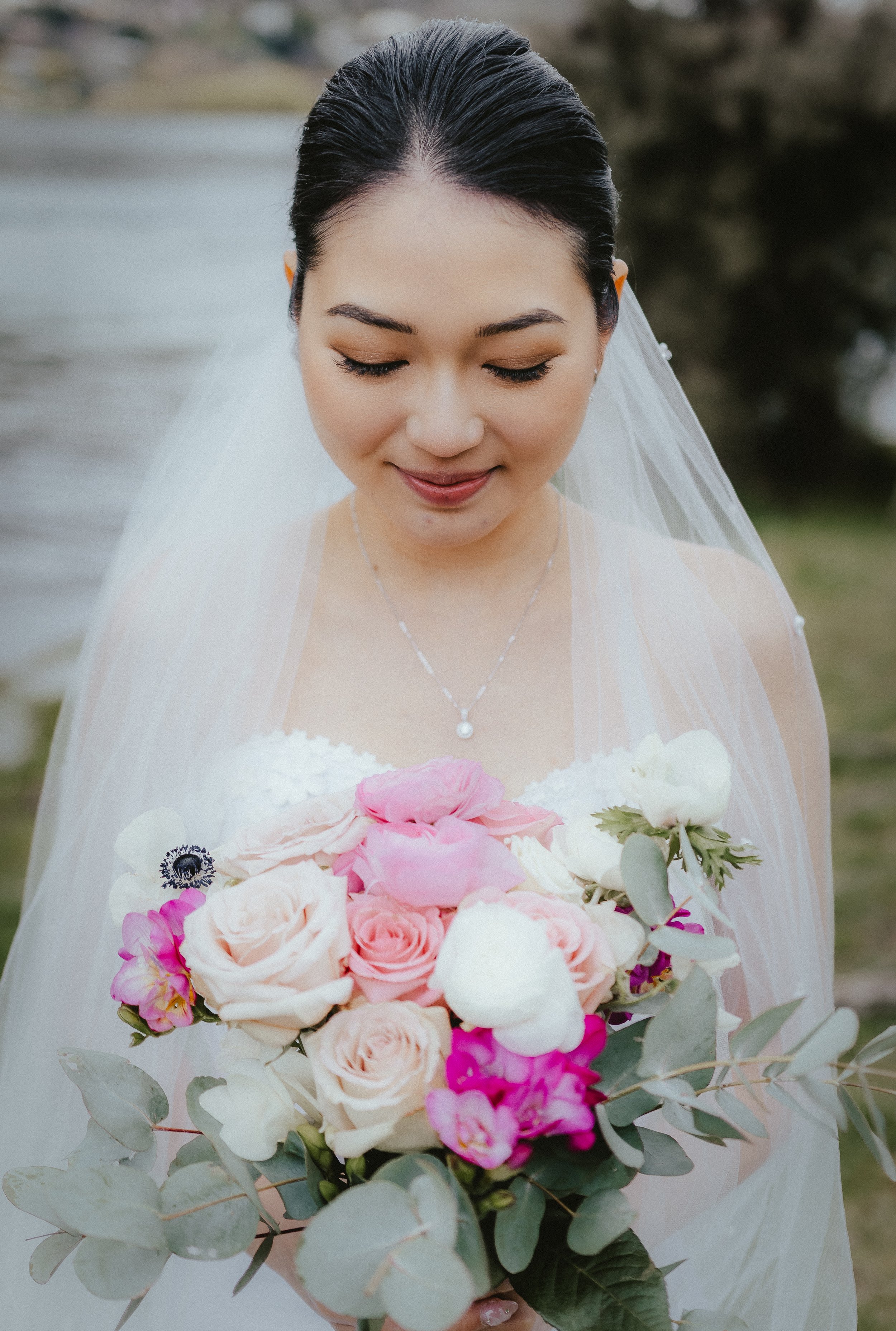 A bride with dark hair styled in an updo, wearing a white wedding dress, holding a bouquet of pink and white flowers with greenery and eucalyptus, and smiling with her eyes closed outdoors.