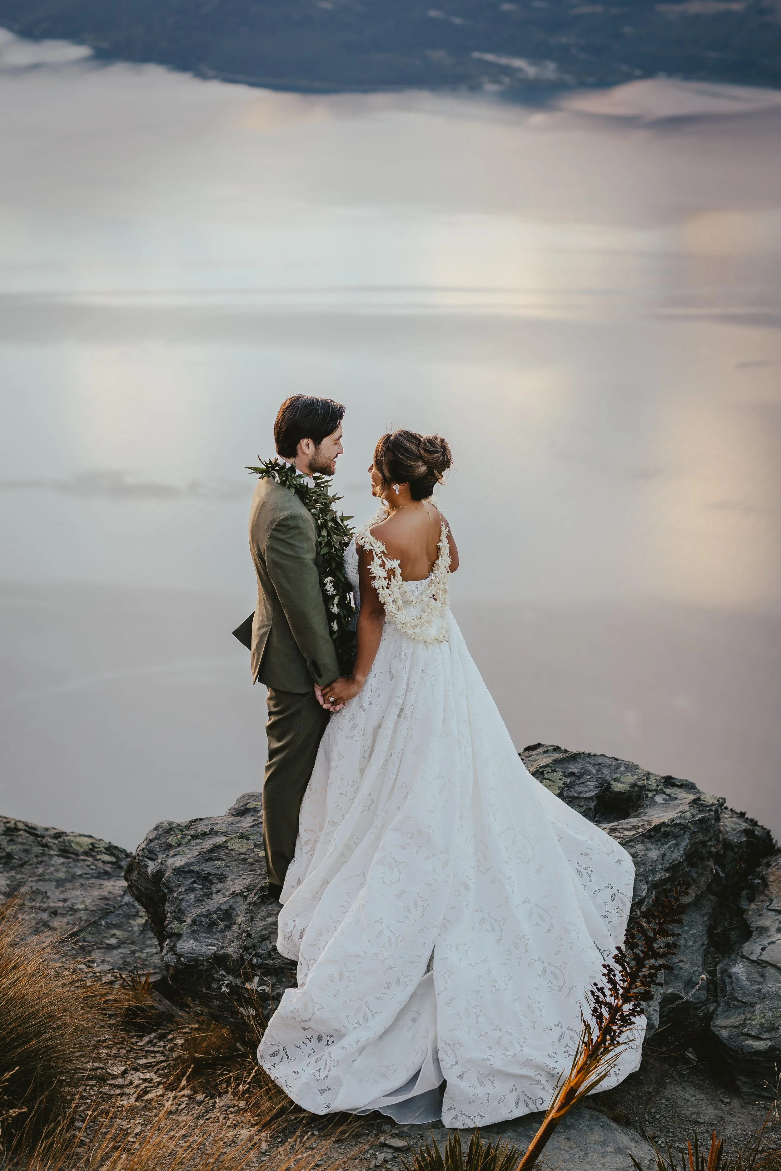 A bride and groom standing on rocks by the water, facing each other and holding hands, during a sunset or sunrise with a cloudy sky in the background.