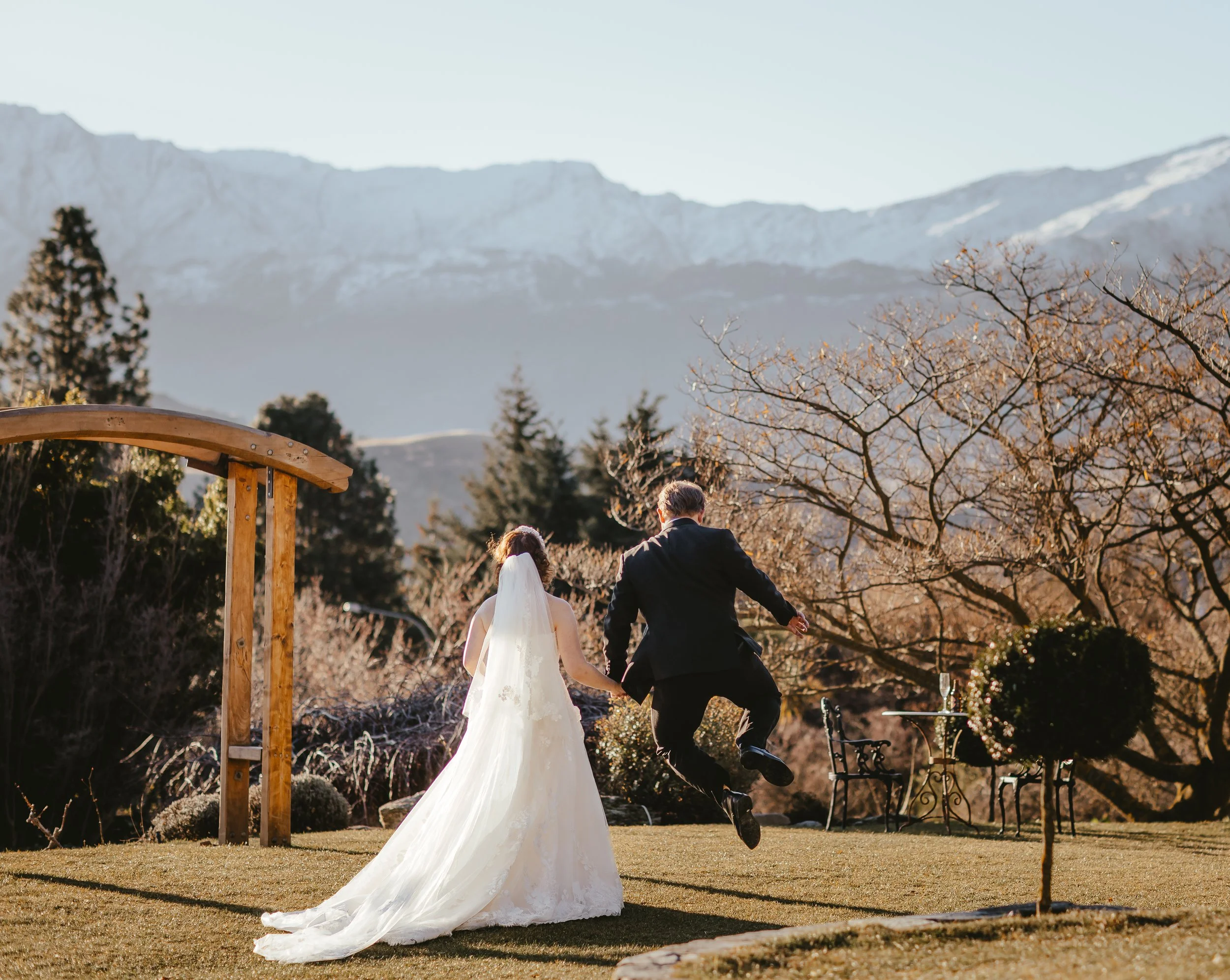 A wedding couple, holding hands, walks away from the camera outdoors in a scenic landscape with mountains, trees, and a clear sky.