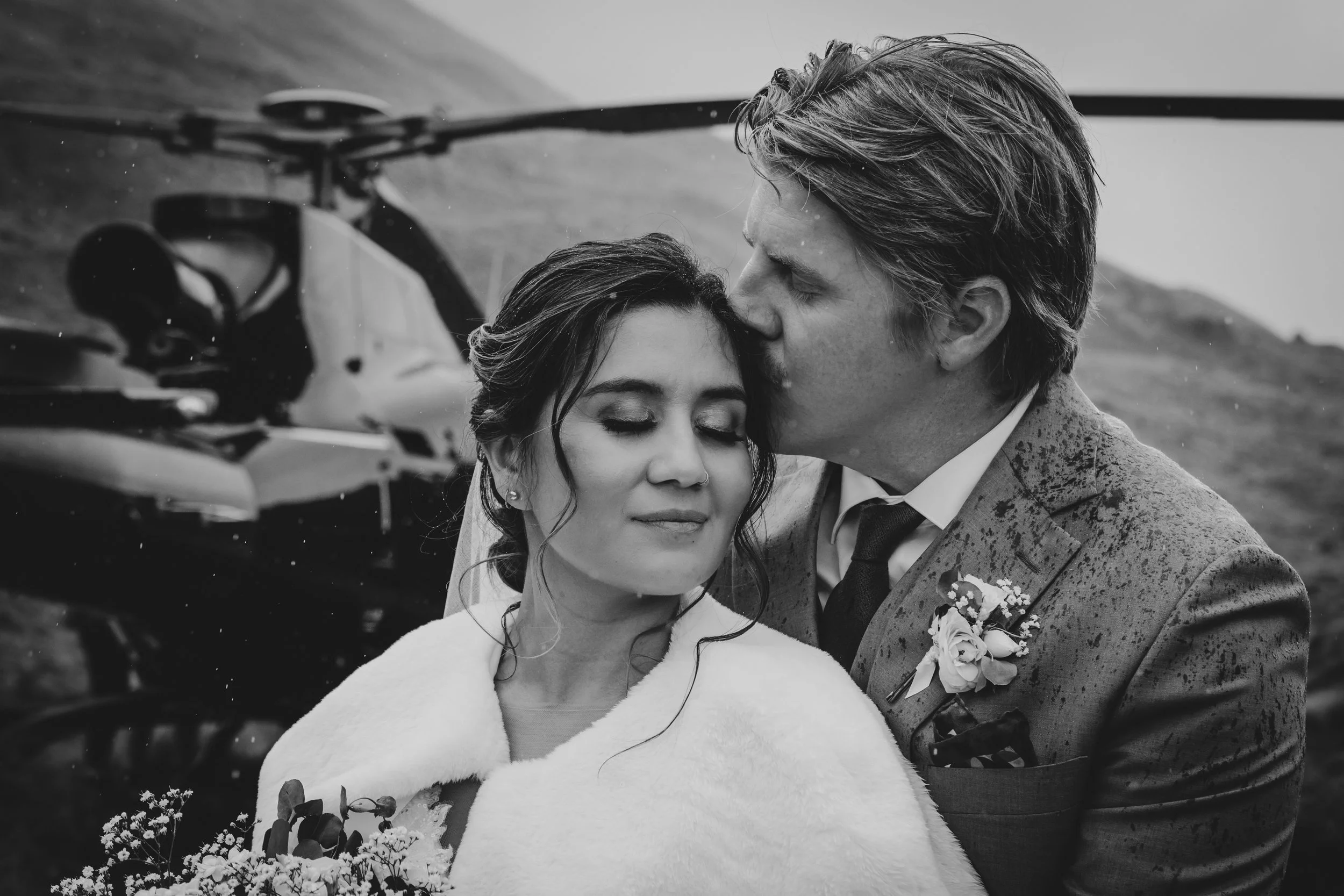 Black and white photograph of a bride and groom sharing a kiss outdoors. The bride has her eyes closed and is holding a bouquet, wearing a white dress and a faux fur shawl. The groom, with disheveled hair, wears a suit with a tie and a boutonniere. A