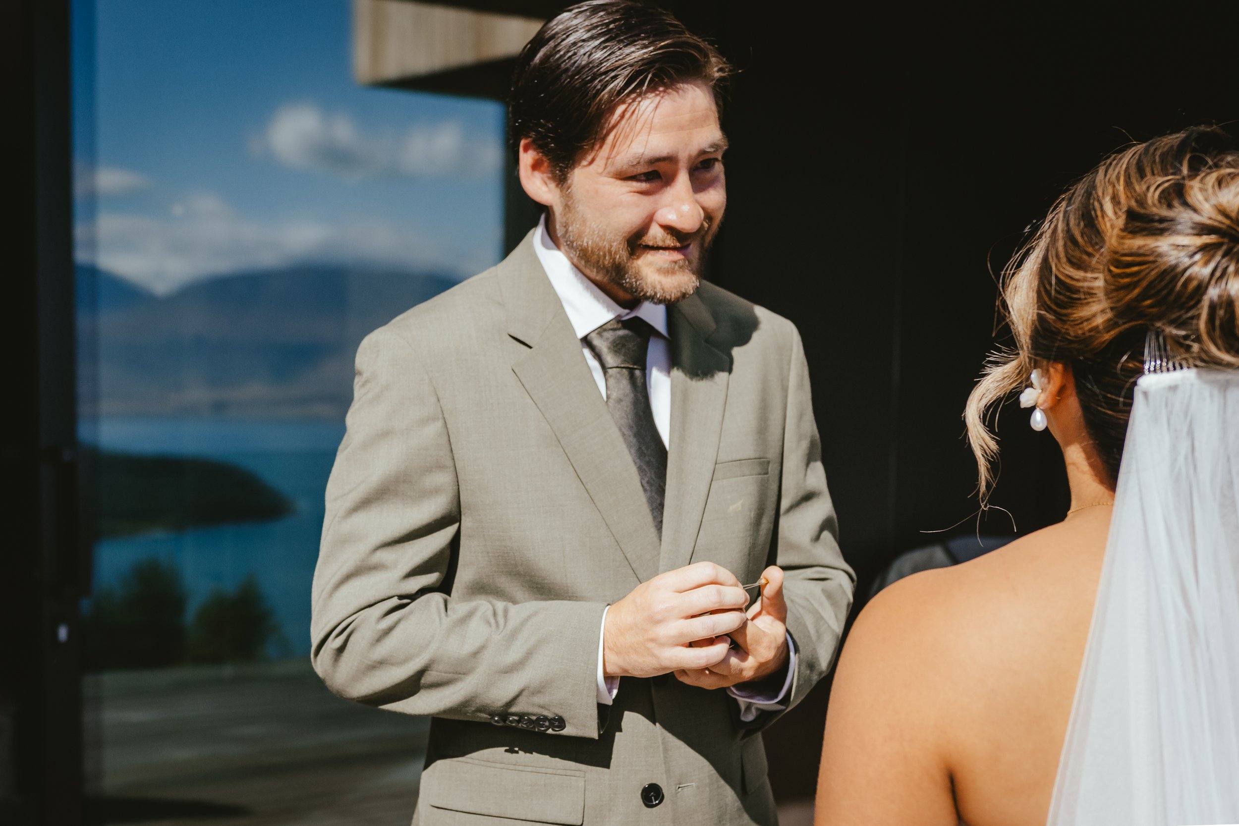 A man with dark hair and a beard in a gray suit is smiling and holding a small object, standing in front of a woman with light hair styled in an updo and wearing pearl earrings, during a wedding ceremony.
