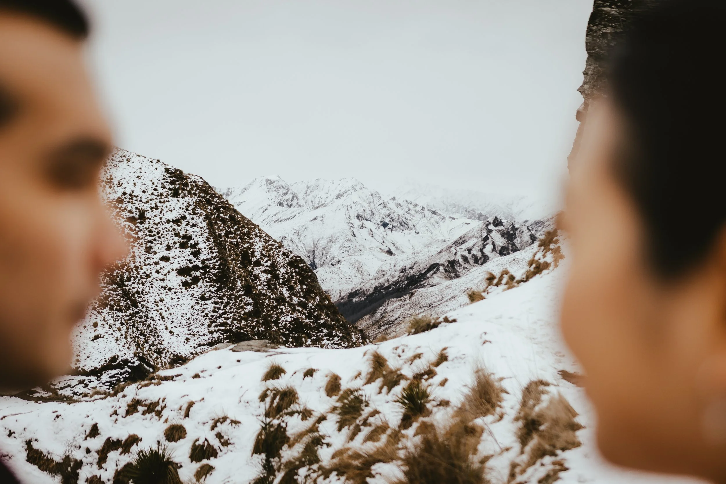 Snow-covered mountains and valleys viewed through a gap between two blurred foreground objects, possibly people's shoulders or heads.