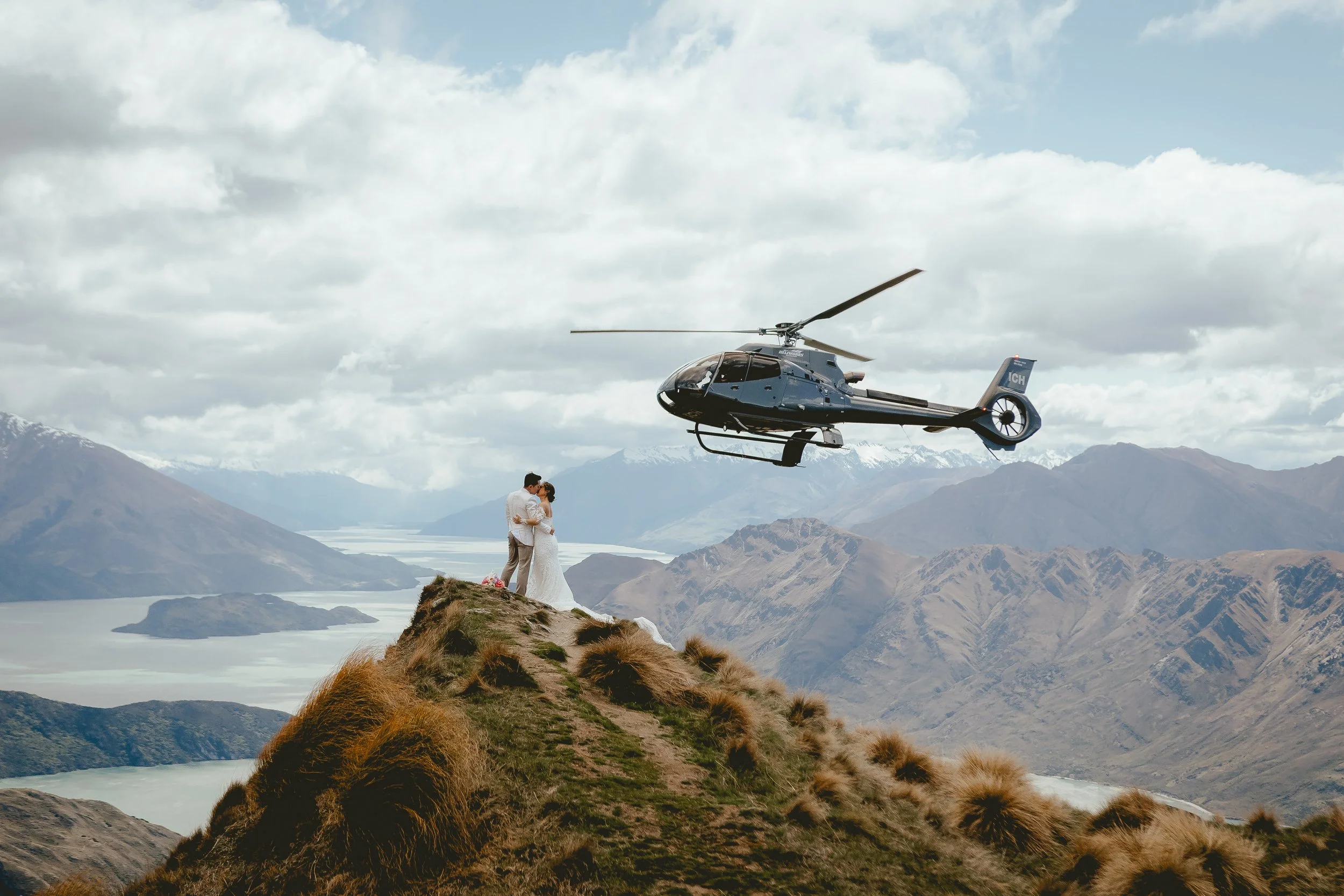 A helicopter flying in the sky over a mountainous landscape with a couple standing on a hilltop, appearing to embrace.