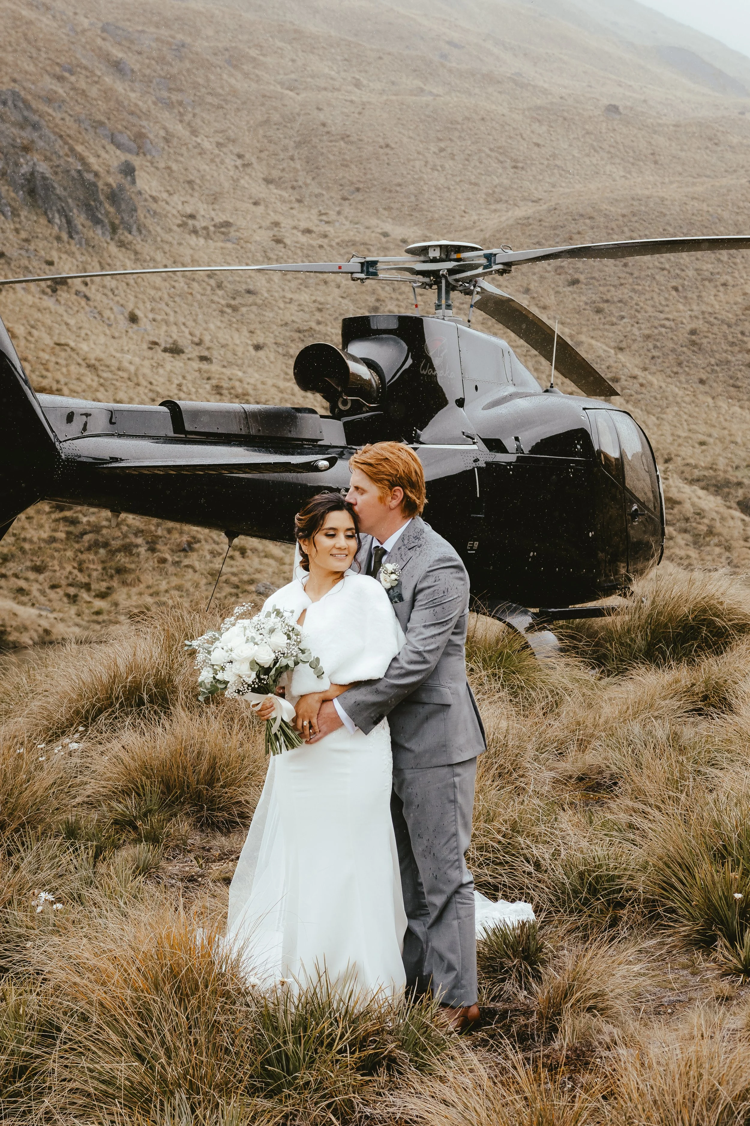 A newlywed couple standing in a grassy field with a helicopter in the background on a mountain hillside.