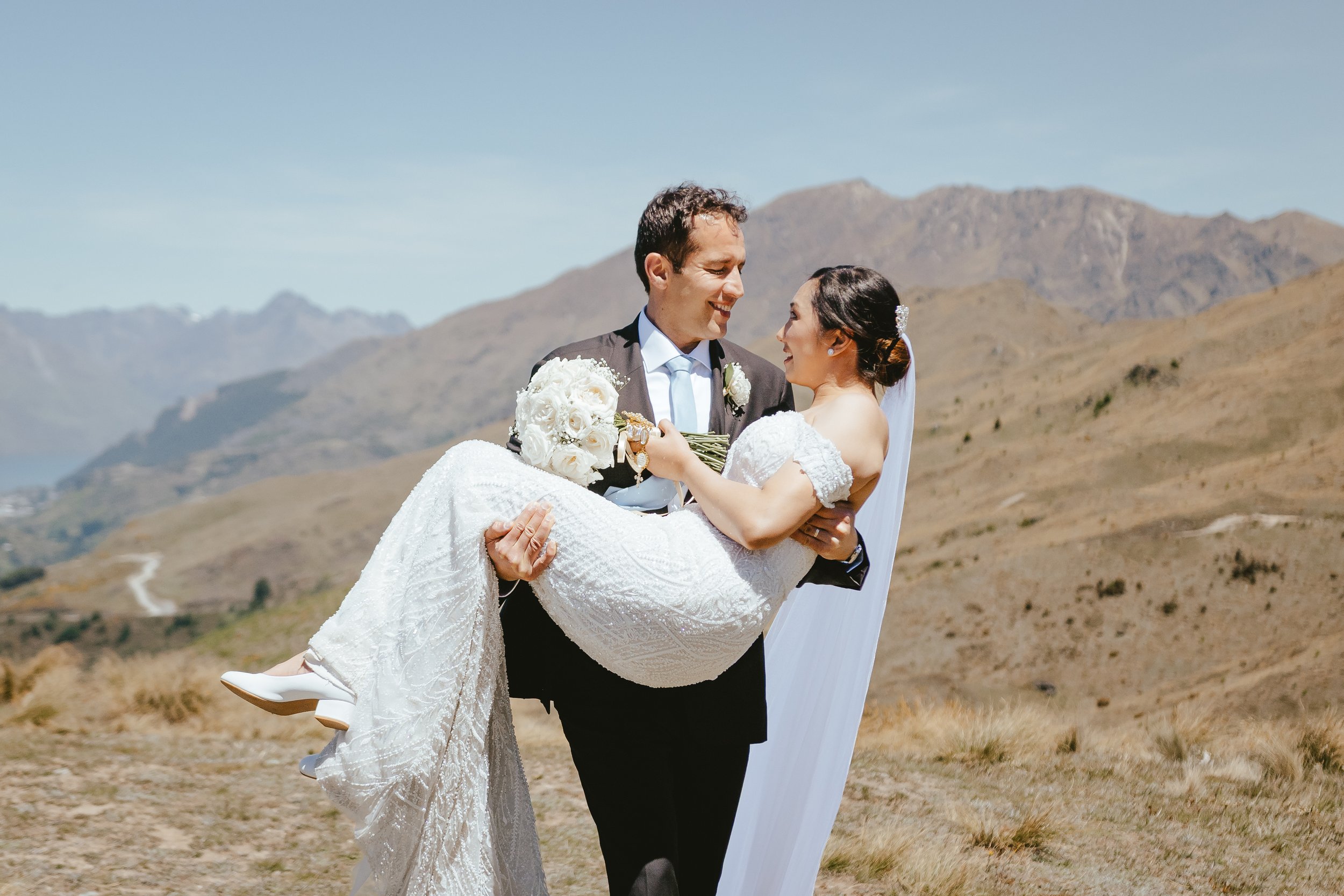 A groom carrying a bride in a white wedding dress across a dry, mountainous landscape during daytime.