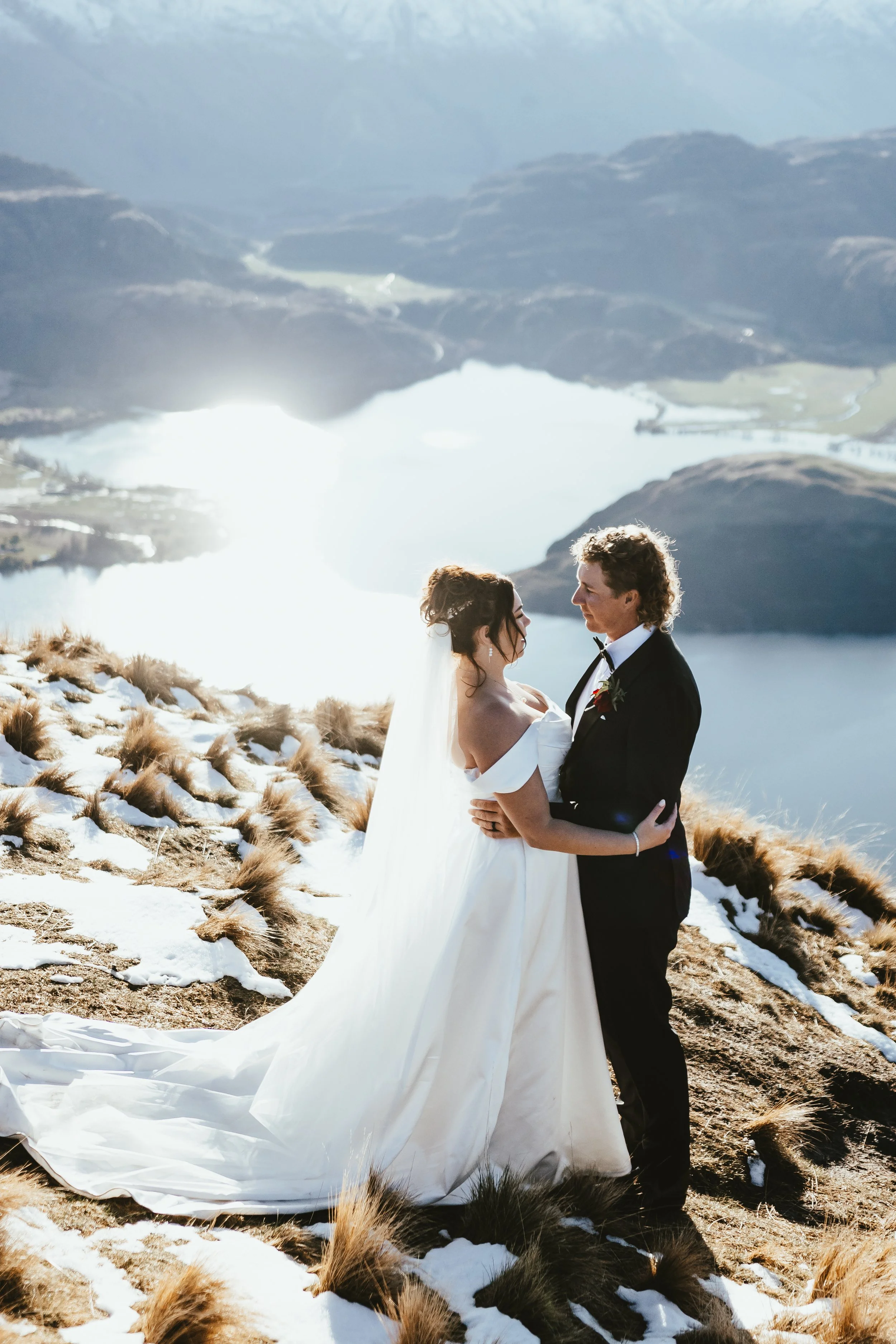 A bride and groom in wedding attire embracing each other outdoors on a snowy and grassy hillside with a lake and mountains in the background.
