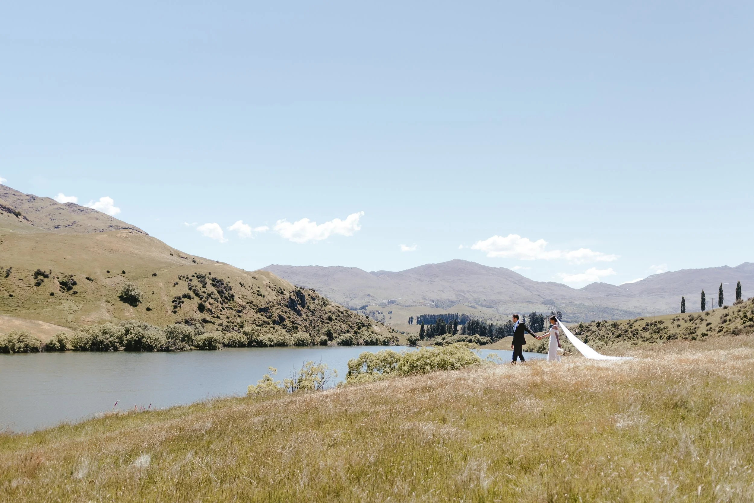 A couple dressed in wedding attire holding hands, walking along a lake with mountains in the background and a partly cloudy sky.