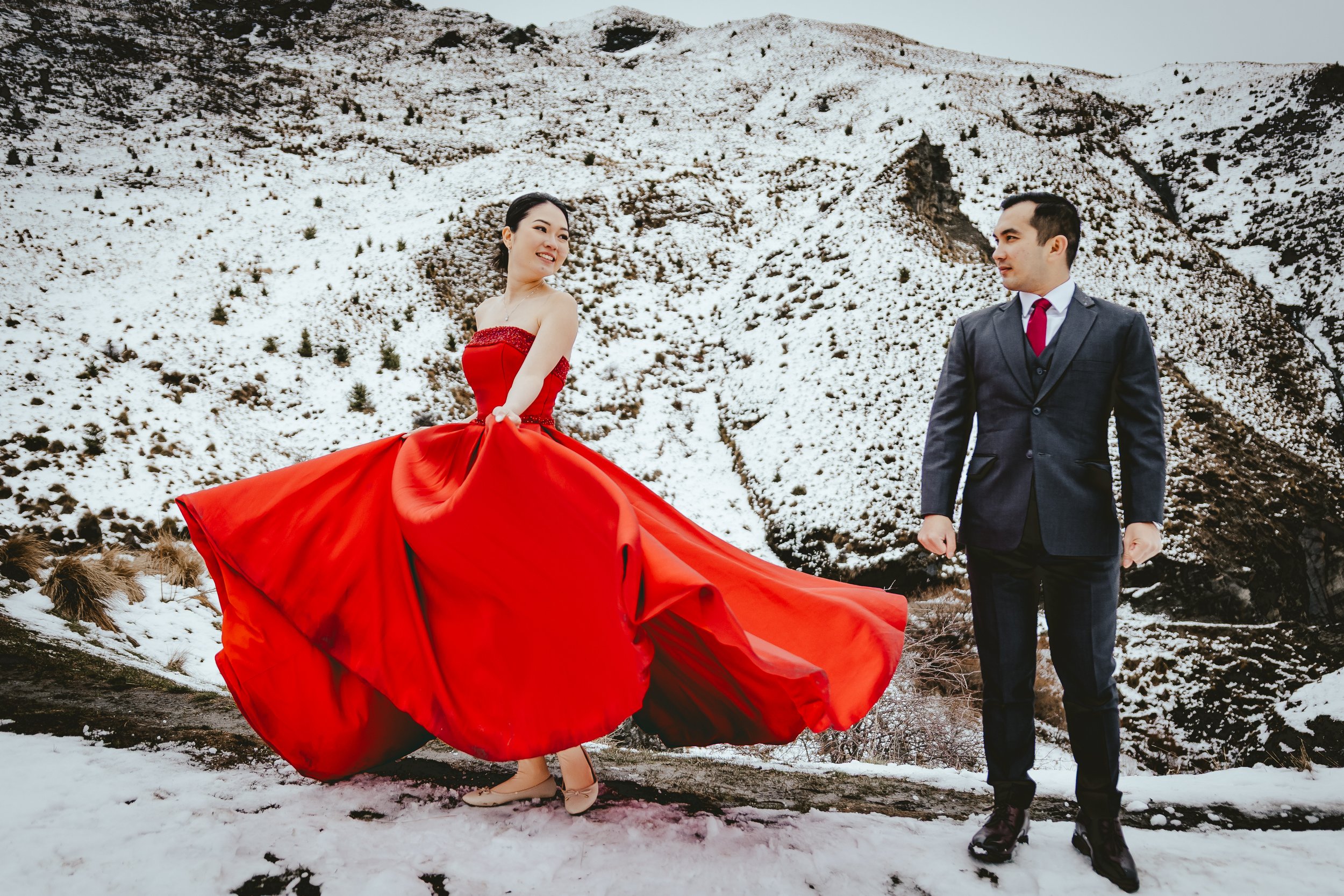 A woman in a red strapless dress and a man in a suit standing in a snowy mountainous landscape.