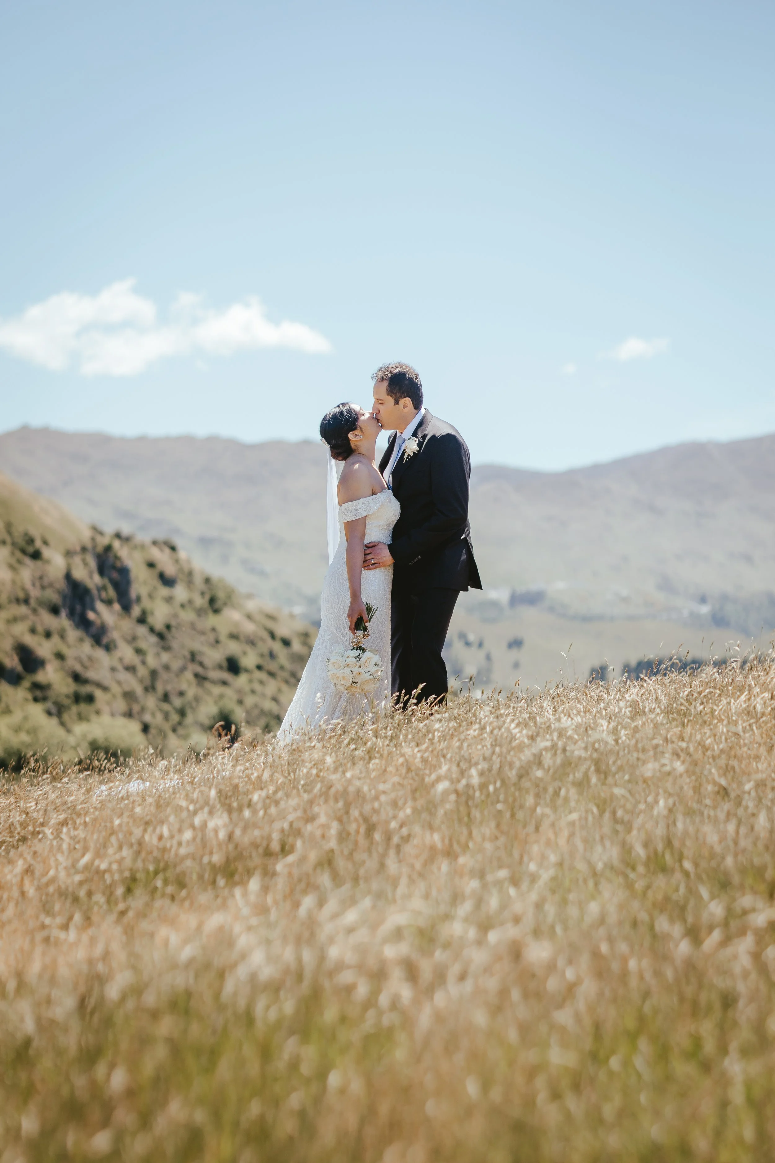 A couple dressed in wedding attire sharing a kiss outdoors in a scenic landscape with mountains and a blue sky.