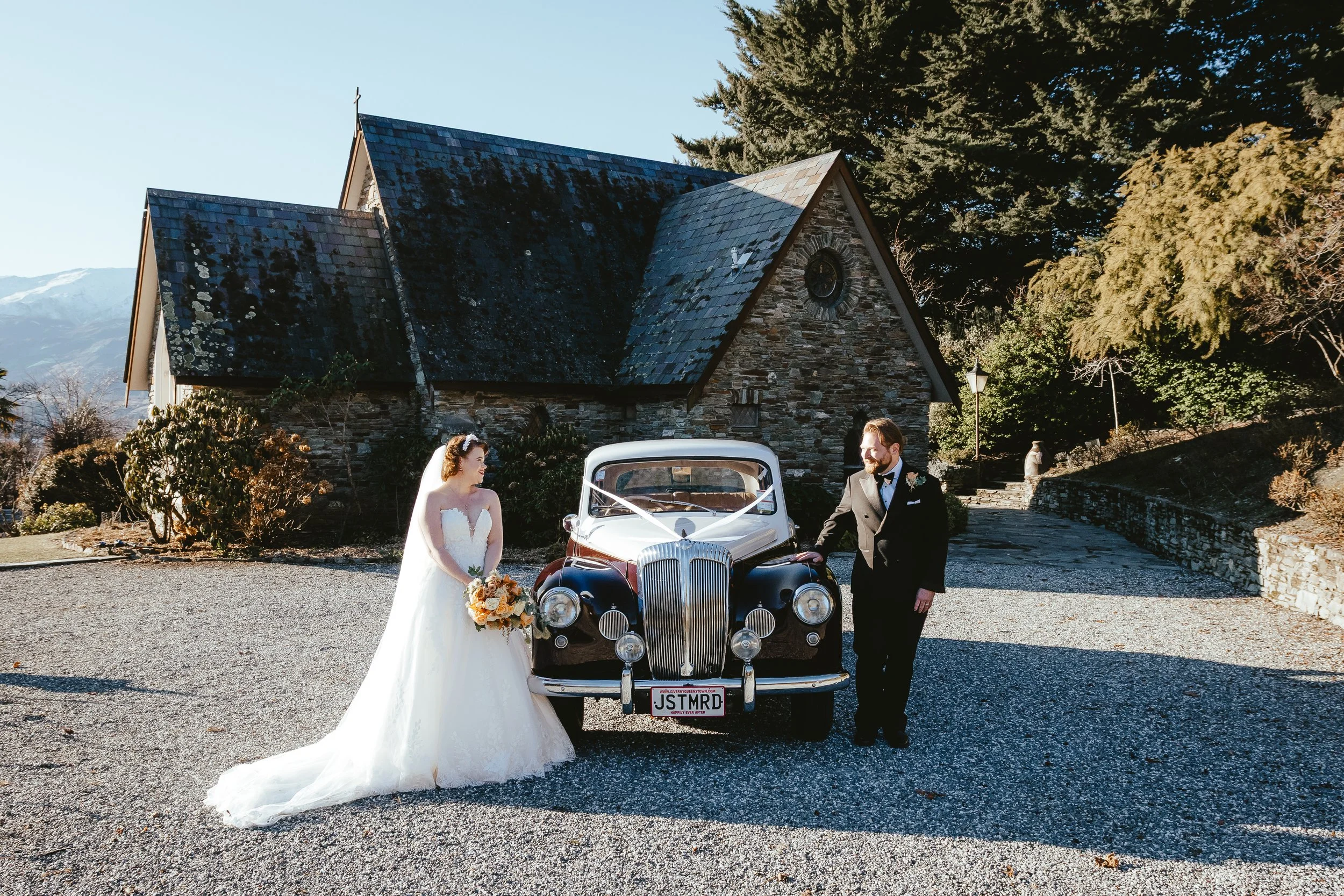 A bride and groom in wedding attire stand beside a vintage car outdoors in front of a stone church, with mountains in the background. The bride holds a bouquet, and they are smiling at each other.