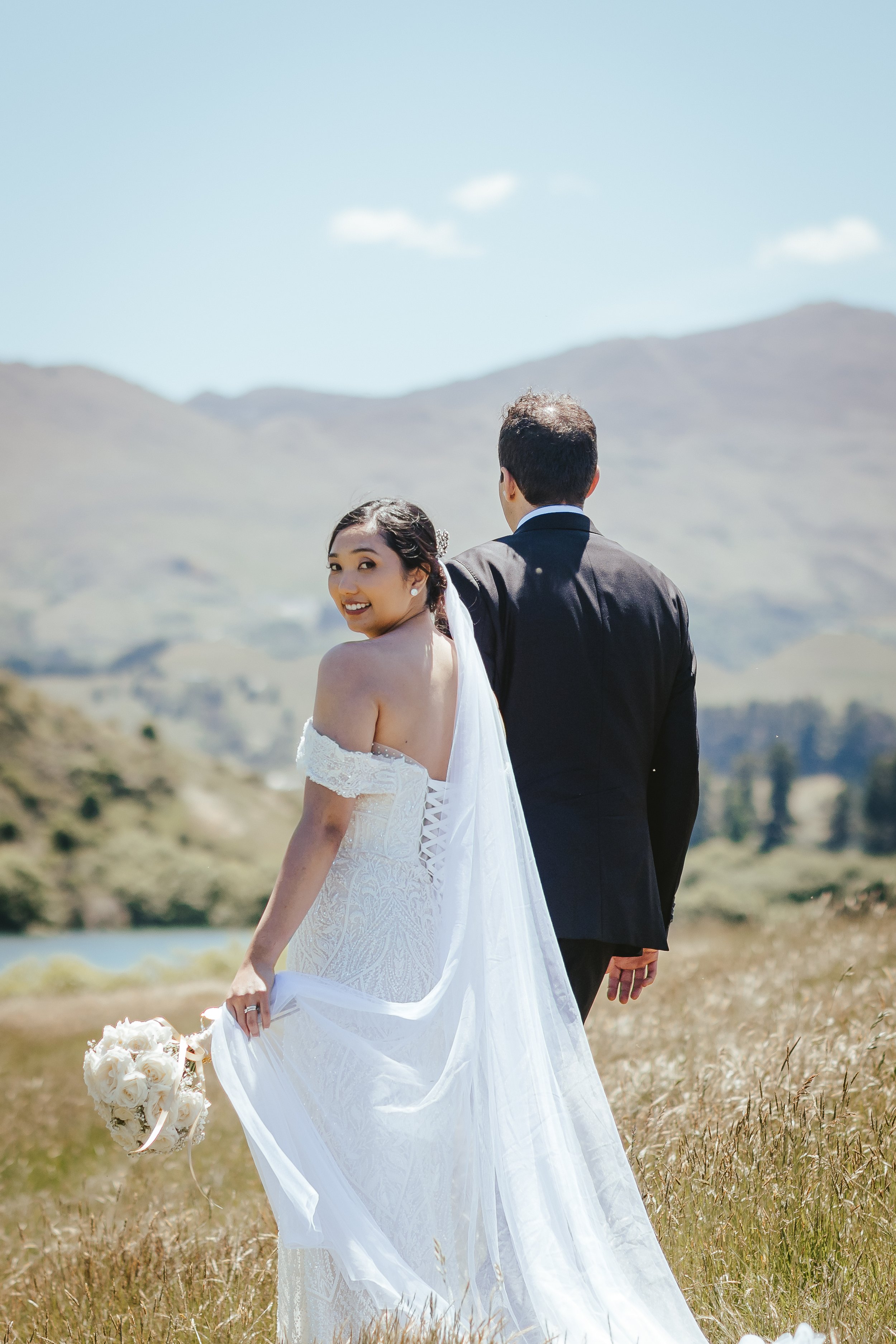 A bride in a white lace wedding dress holding a bouquet of white roses, smiling while looking over her shoulder. A groom in a black suit stands nearby, facing away. They are outdoors on a sunny day near water with mountains in the background.
