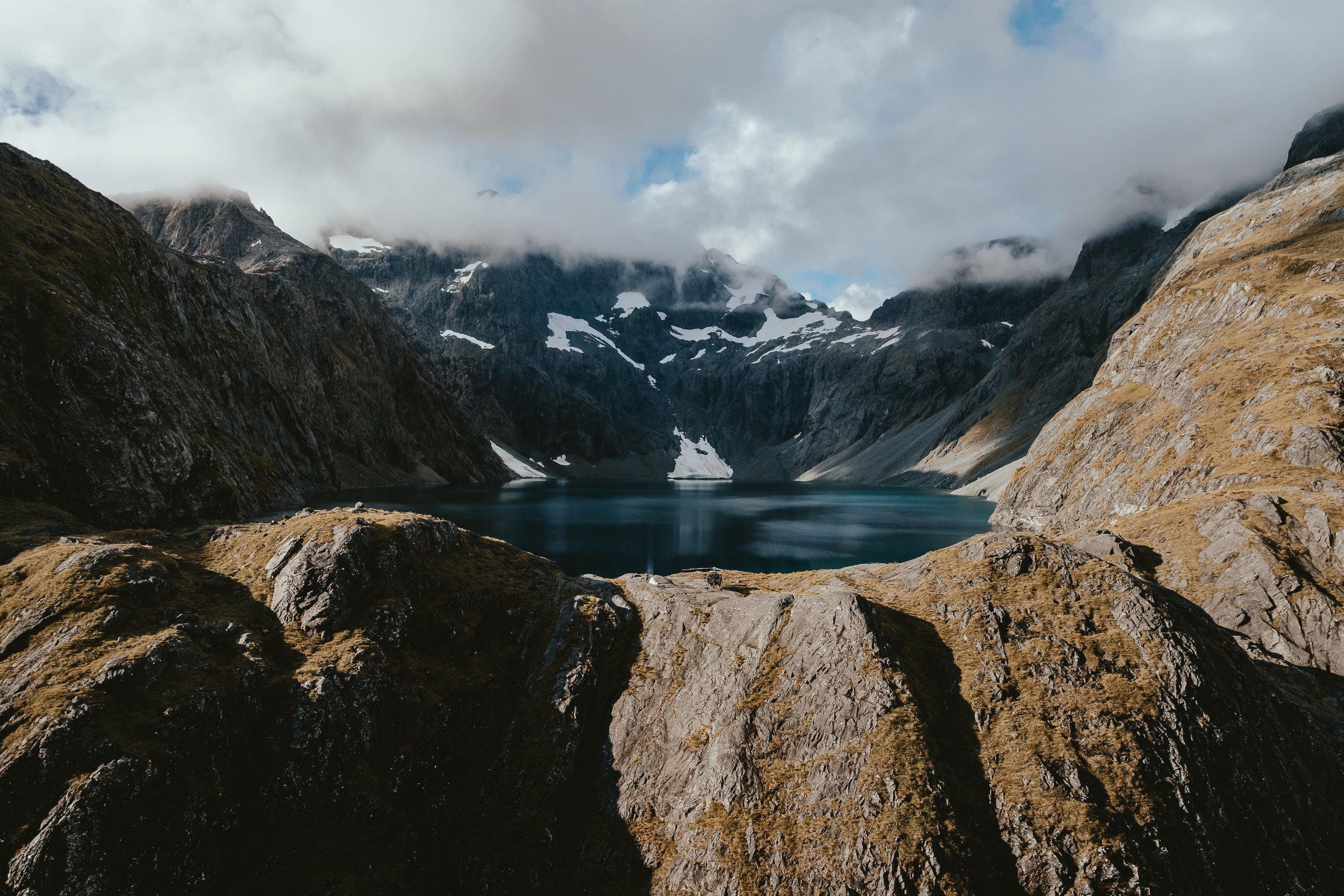 Mountain landscape with rocky cliffs, snow patches, and a lake in the foreground, partly obscured by clouds.