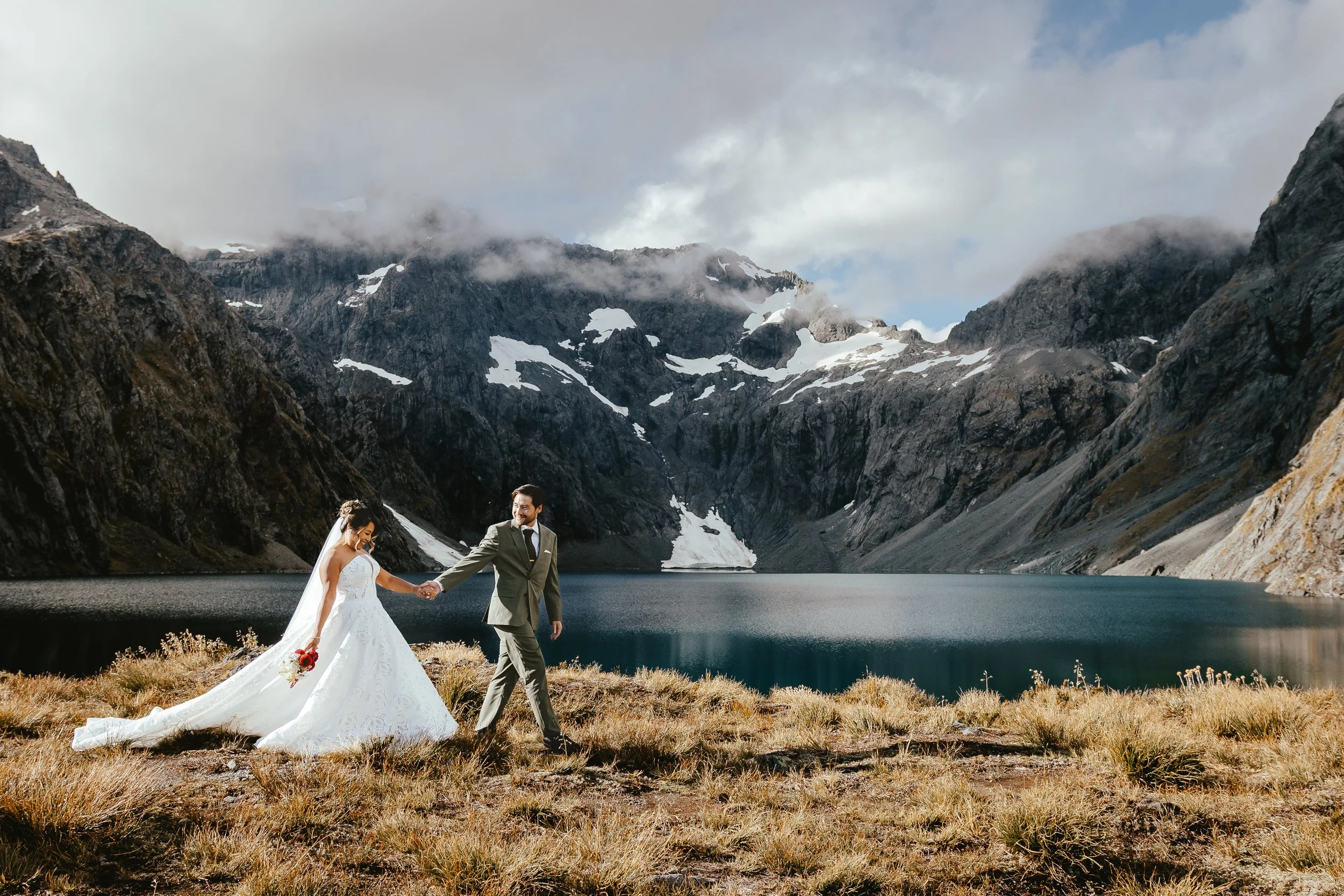 A bride and groom holding hands by a lake surrounded by mountains with snow patches, cloudy sky, and grassy terrain.