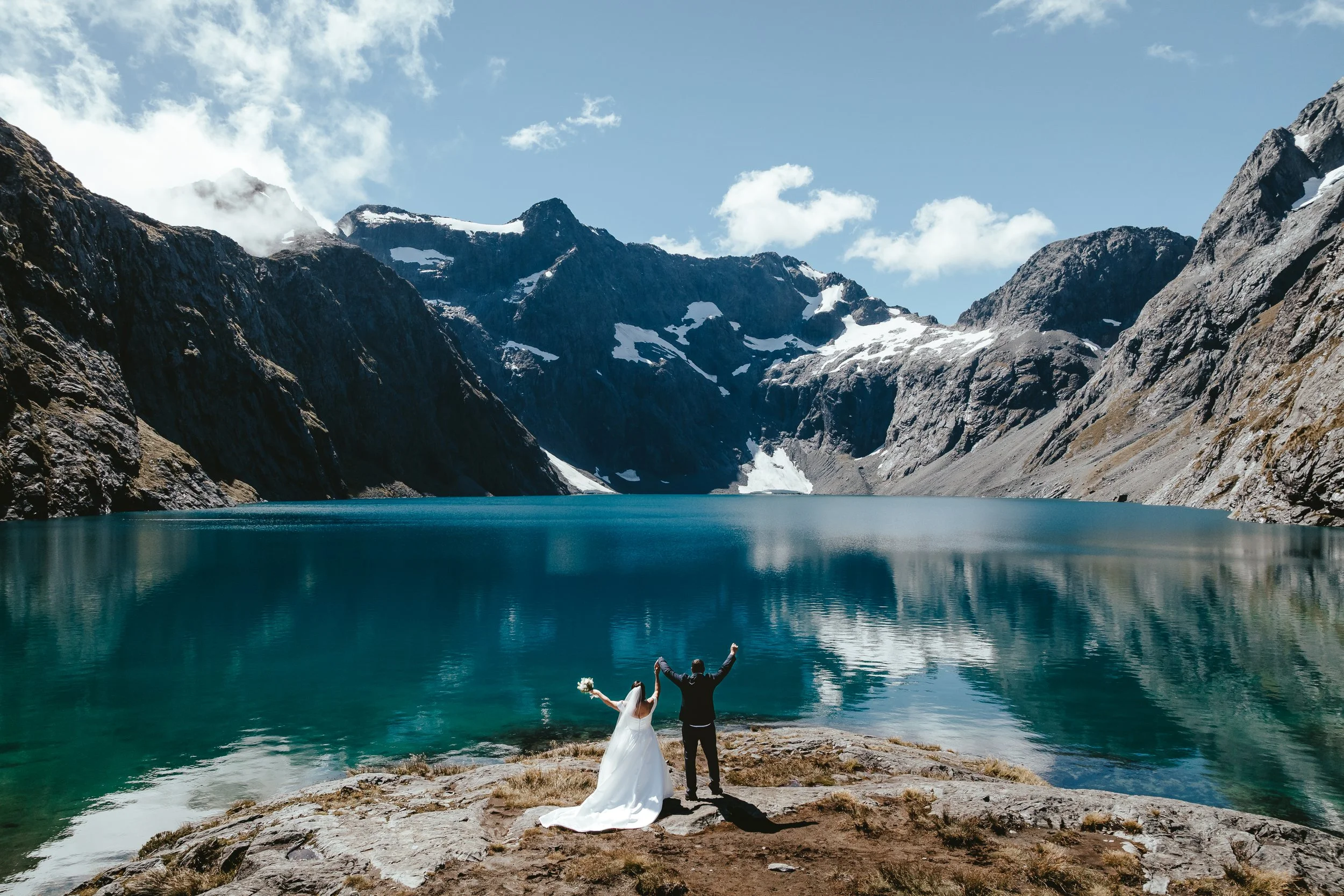 A bride and groom stand on a rocky shore by a large turquoise lake, with snow-capped mountains and a partly cloudy sky in the background, celebrating their wedding.