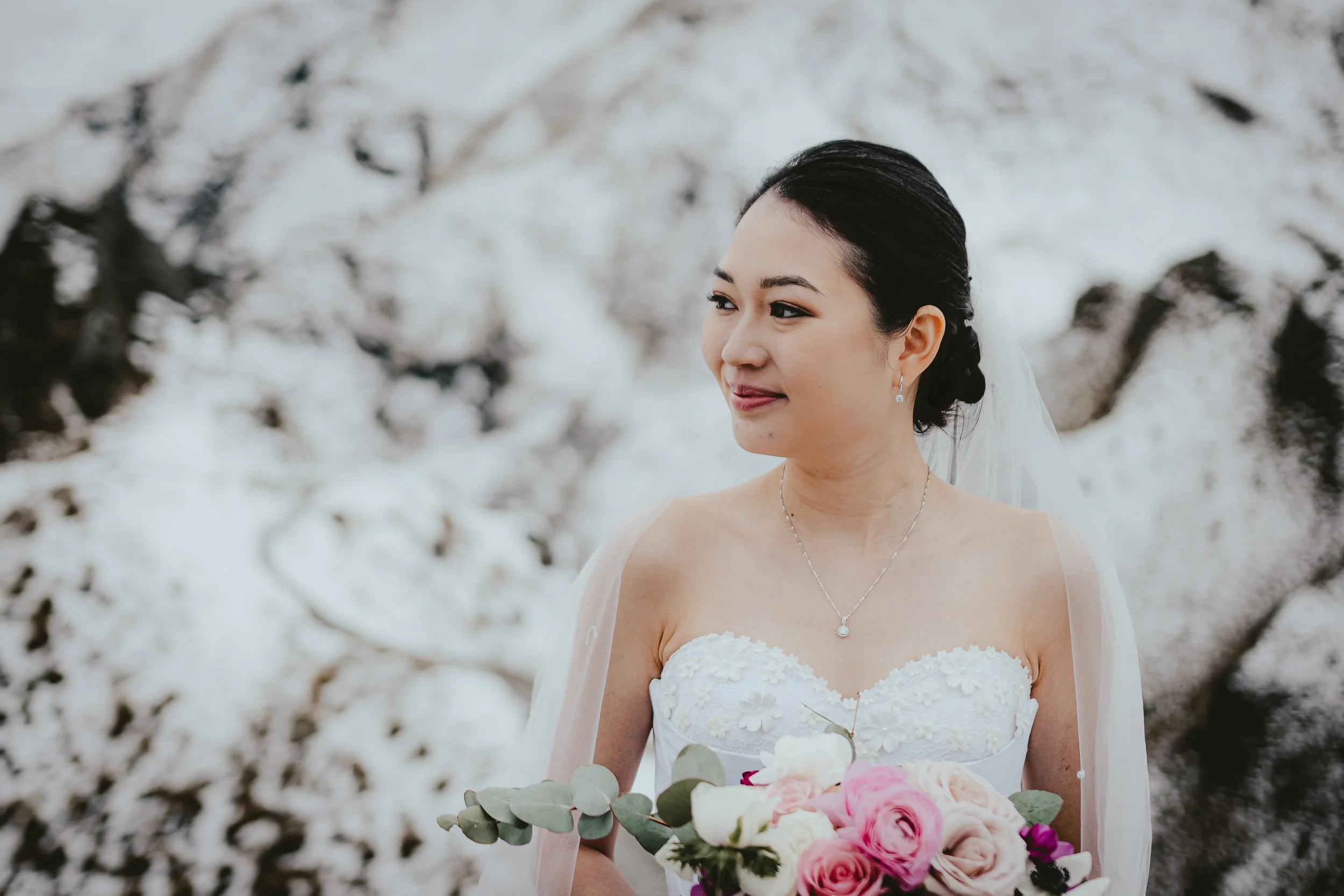 A smiling bride wearing a white strapless wedding dress with floral embroidery, holding a bouquet of pink and white roses with eucalyptus leaves, standing outdoors in a snowy landscape with snow-covered trees in the background.