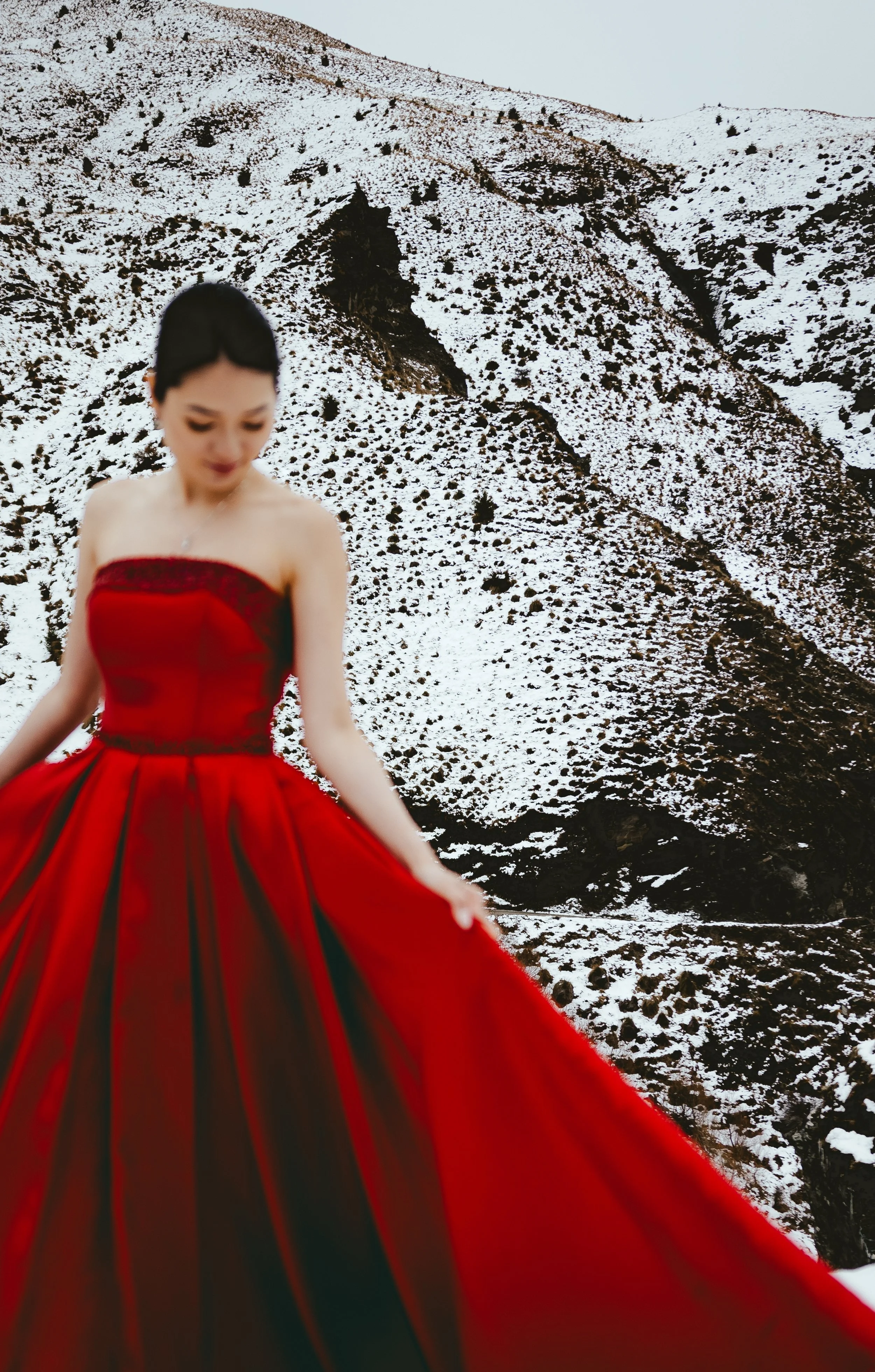 A woman in a strapless red dress with black accents stands in front of snow-covered mountains.