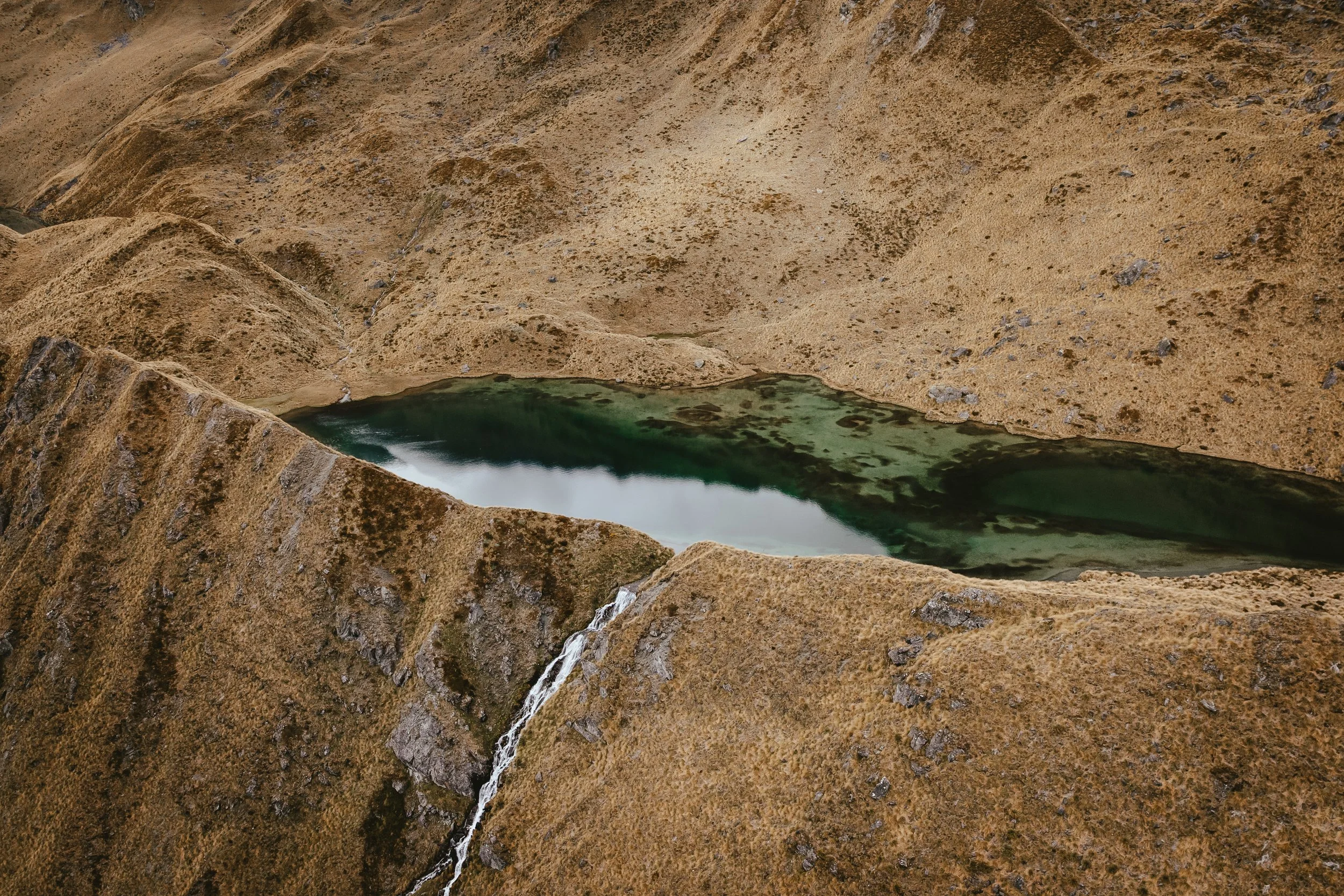 A rugged, barren landscape with sloped, sandy hills surrounding a small body of greenish water.