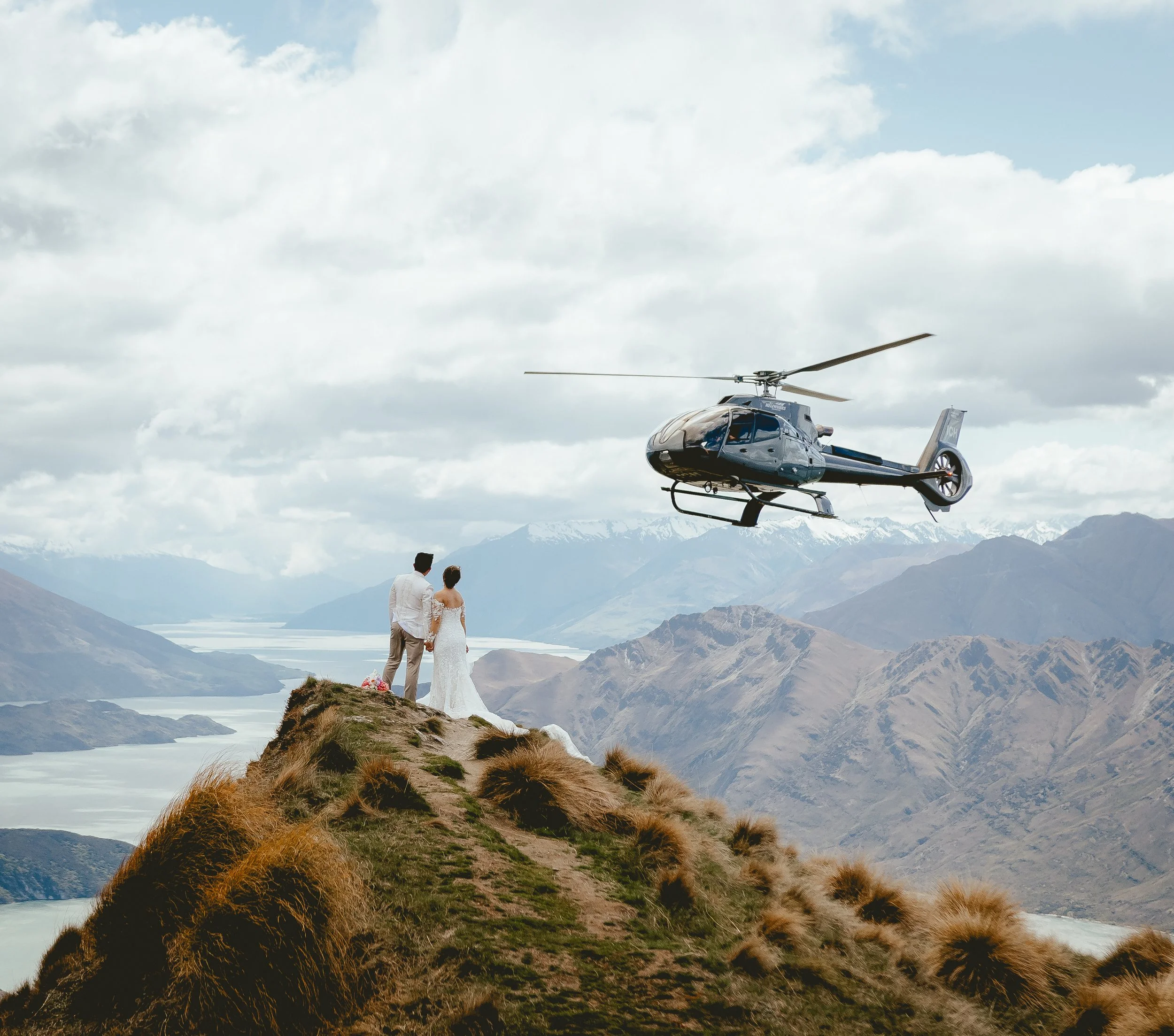 Couple standing on a remote alpine peak during a heli elopement as a helicopter flies past, surrounded by quiet mountain scenery.