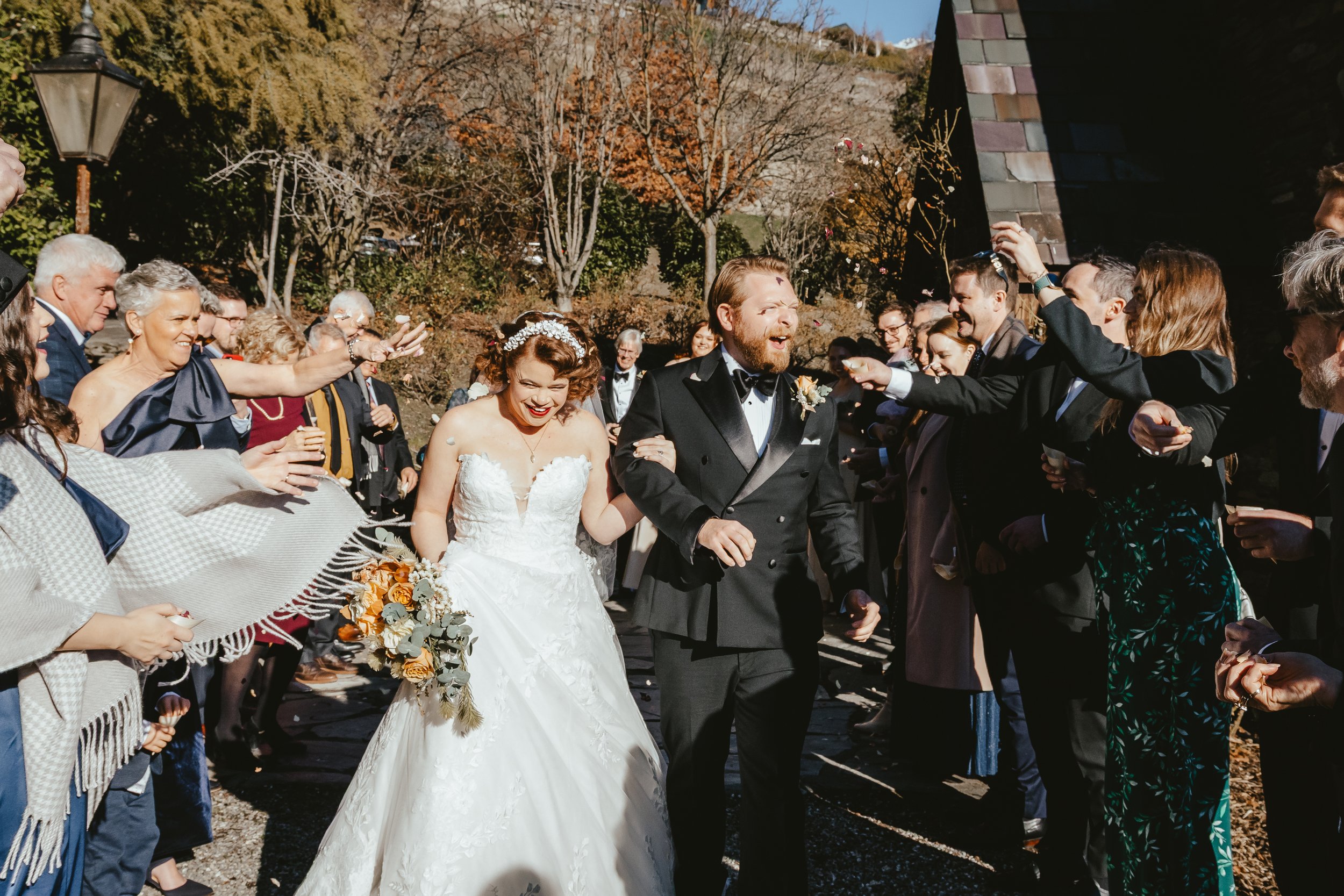 A bride and groom walking together and holding hands at their outdoor wedding, surrounded by smiling guests under a clear blue sky.