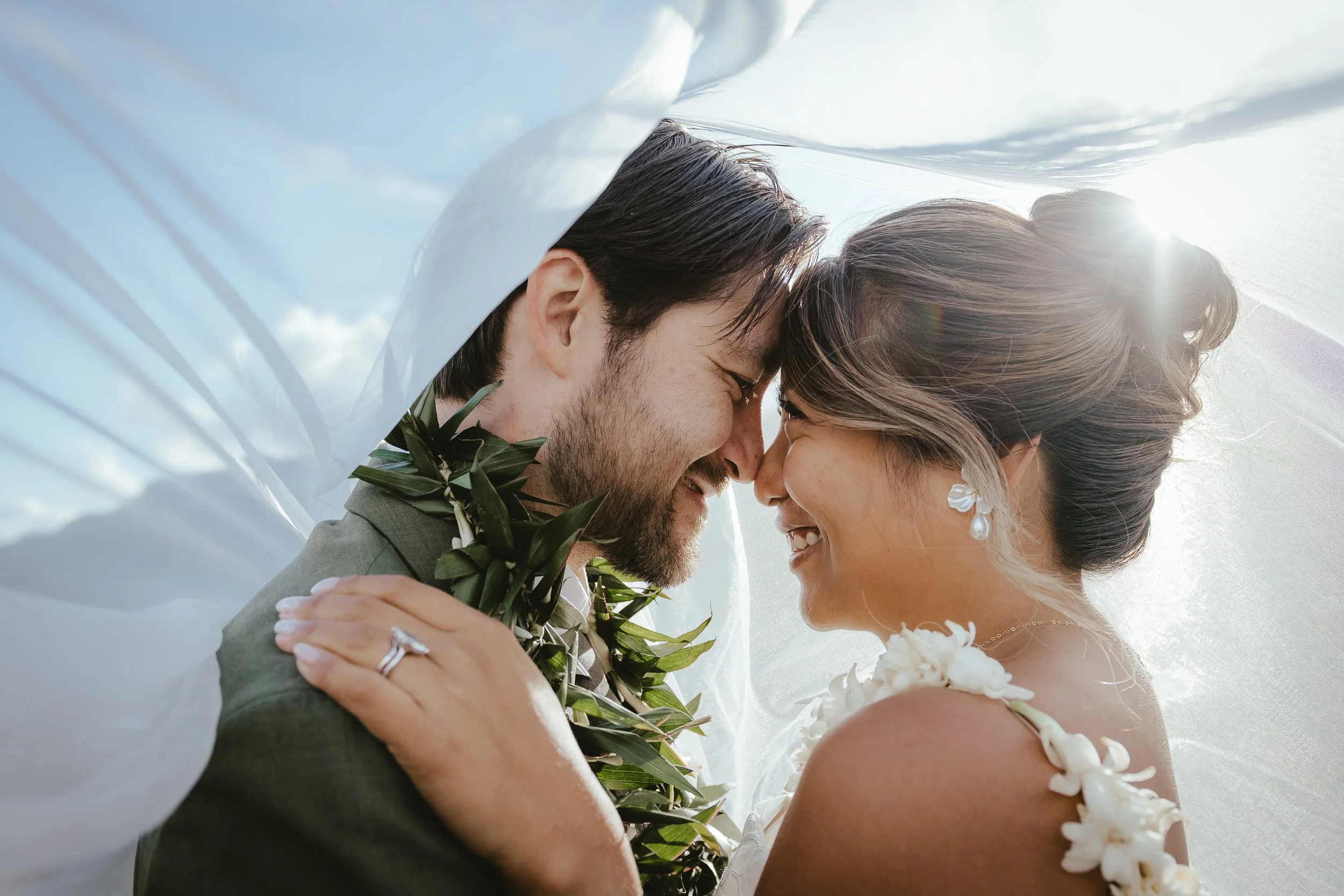 A couple on their wedding day, smiling and touching foreheads, partially surrounded by sheer white fabric with the sun shining behind them.