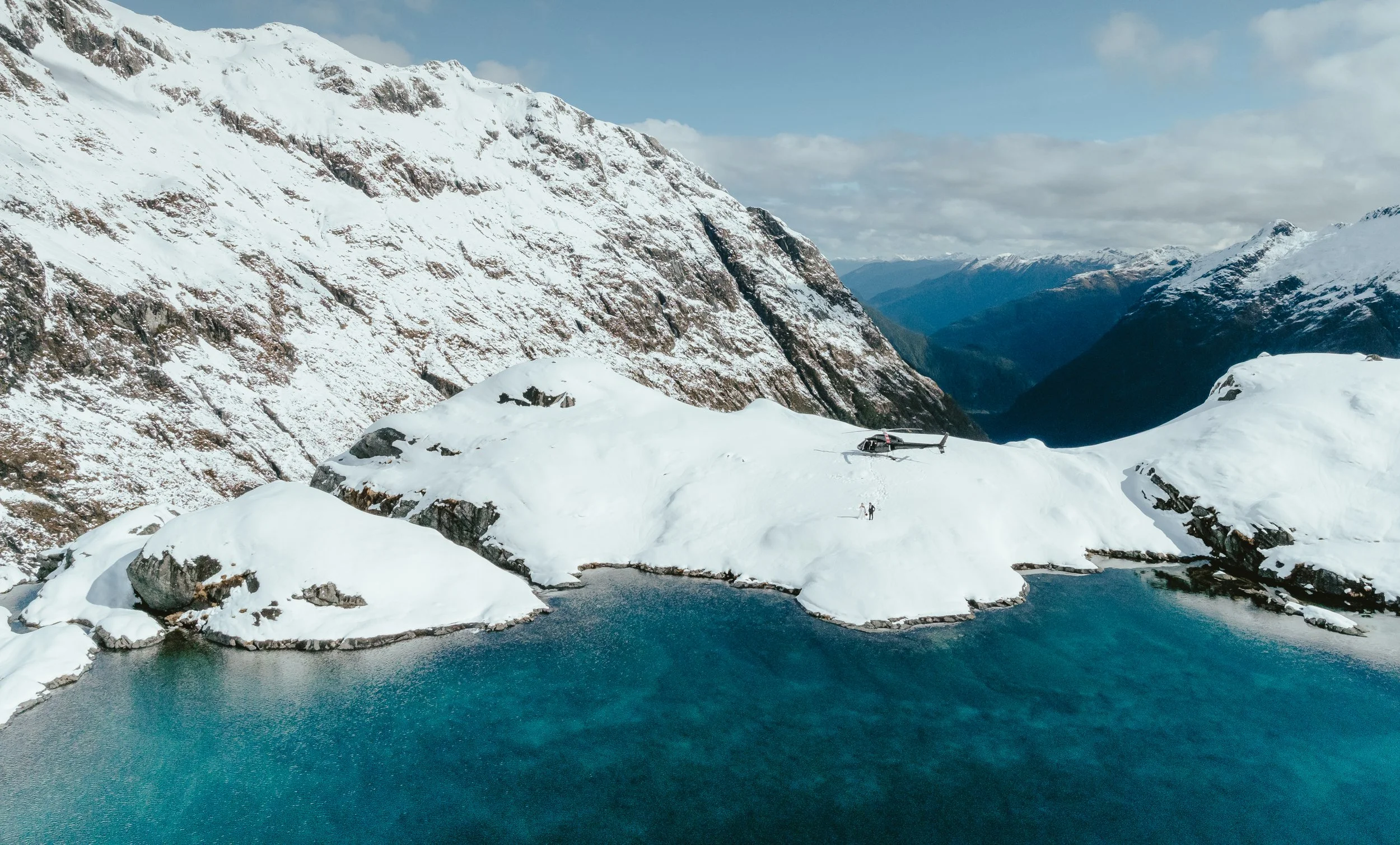 Snow-covered mountains with a helicopter and people on the snow, beside a small icy lake in a remote alpine area.
