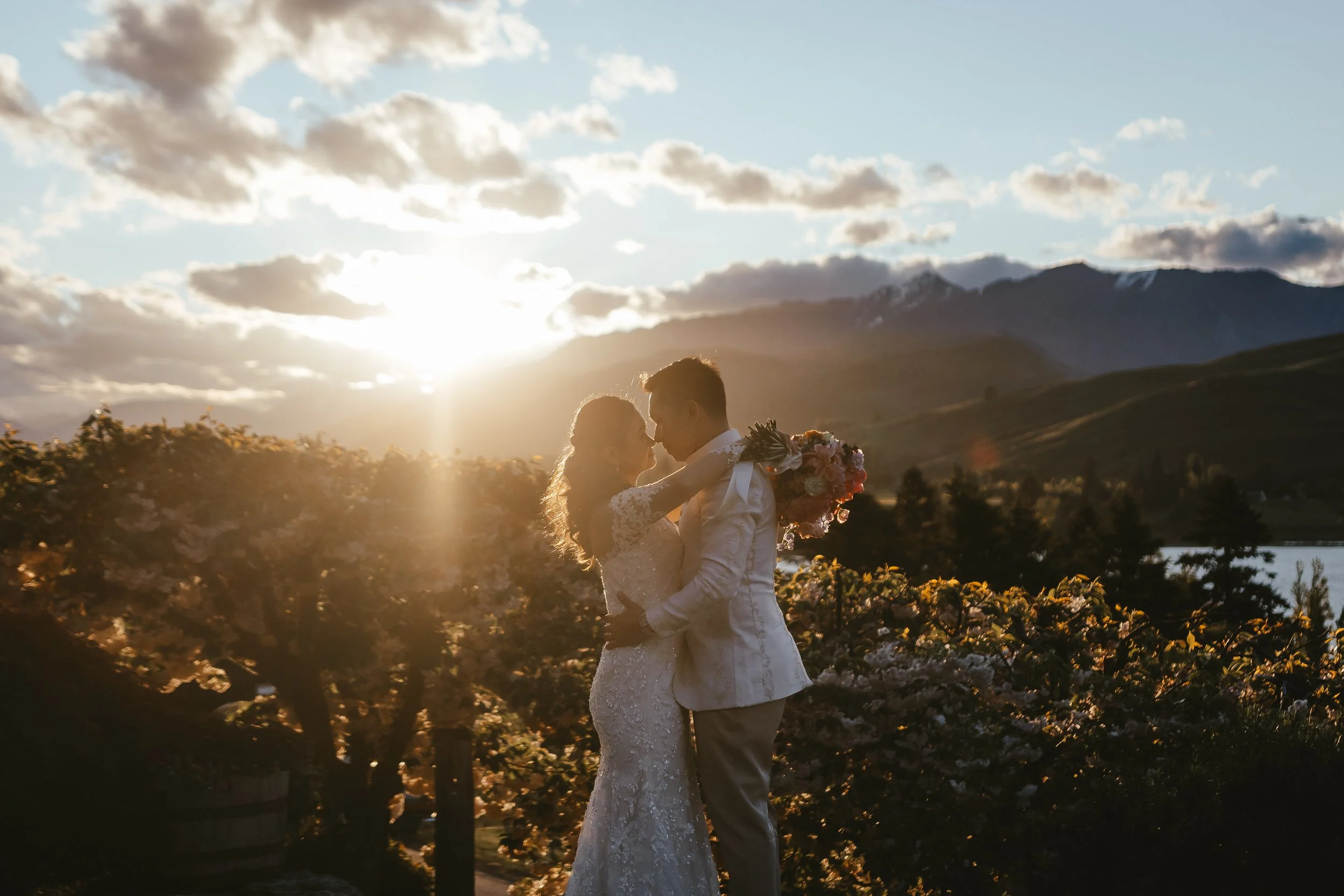 A couple in wedding attire sharing a romantic moment outdoors during sunset, with mountains and a partly cloudy sky in the background.