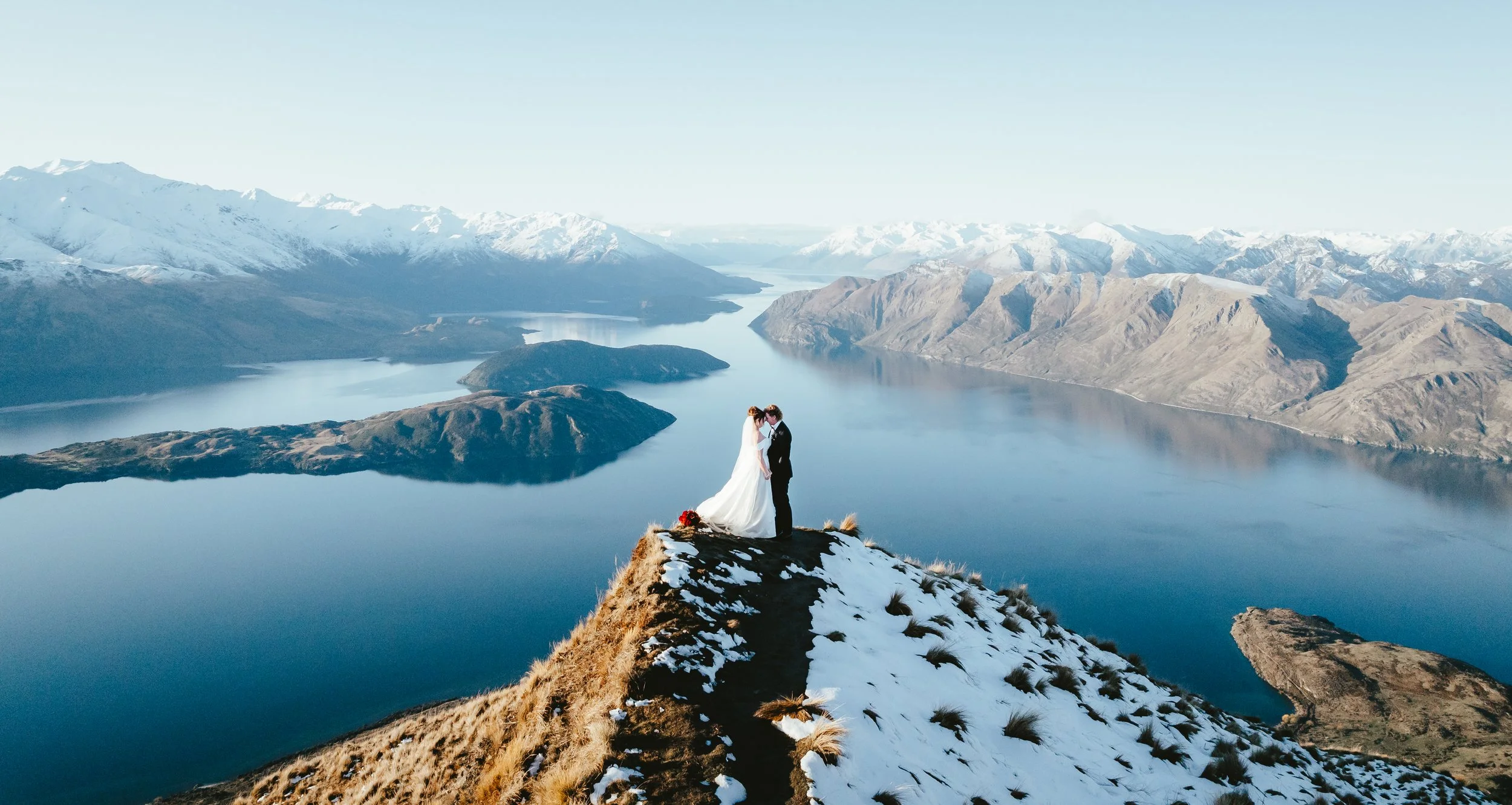 A bride and groom holding hands on a snowy mountain peak with a lake and snow-capped mountains in the background.