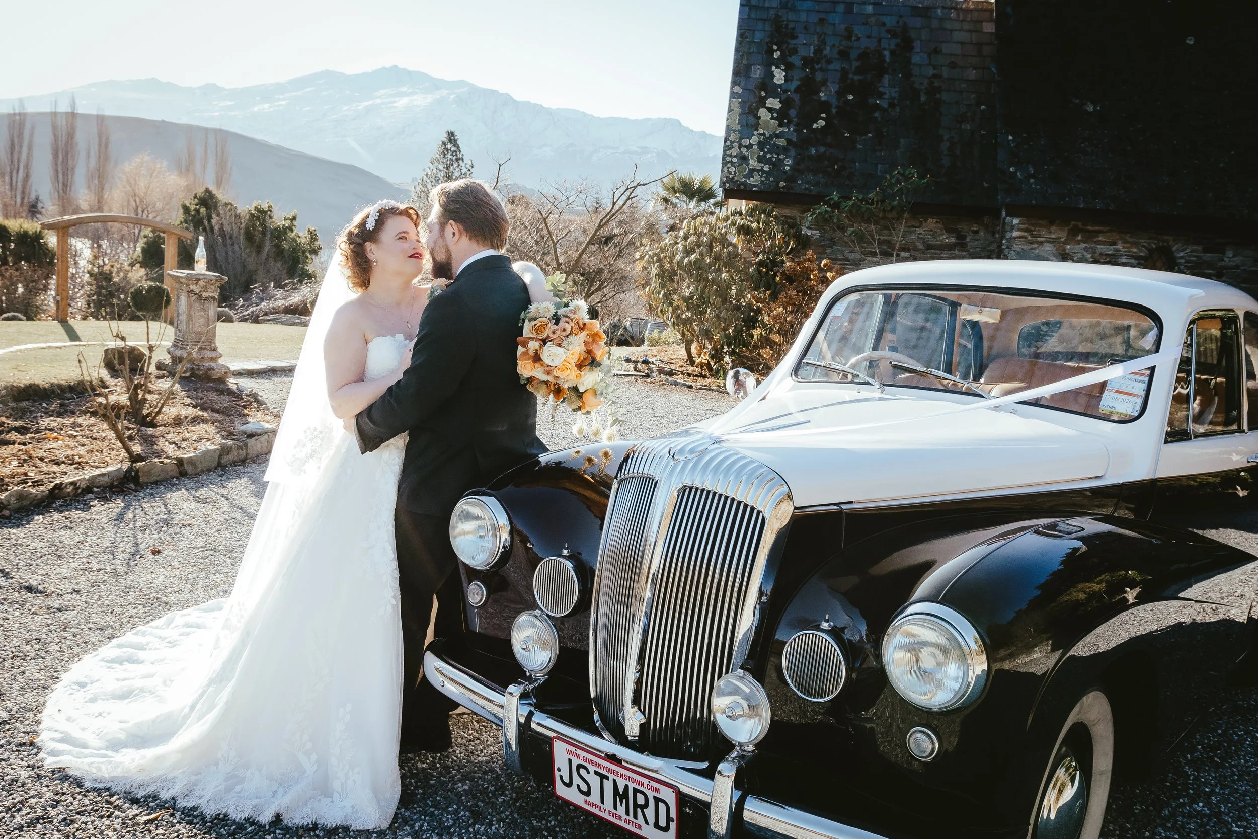 A bride and groom sharing a moment beside a vintage black and white car at an outdoor wedding with mountains in the background.