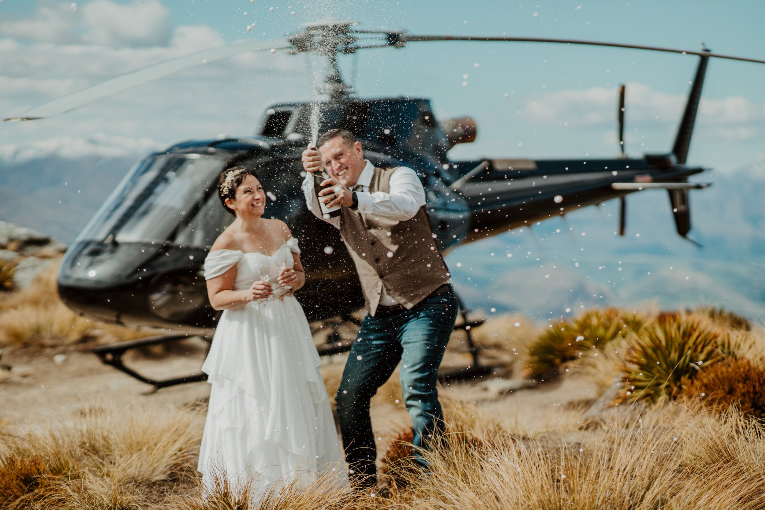 Bride and groom popping champagne in front of their helicopter on Cecil Peak for an unforgettable heli elopement.