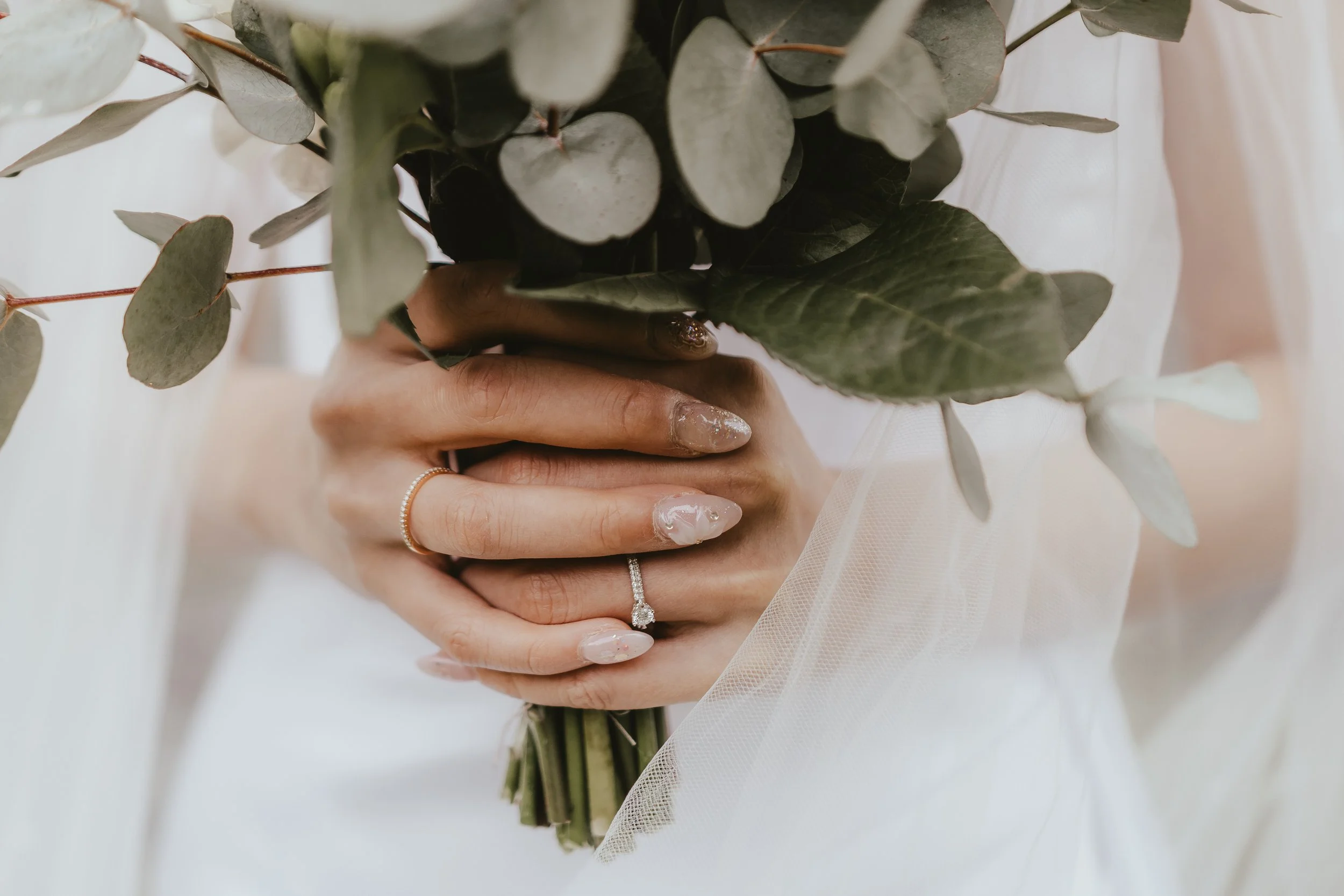 Close-up of a person holding a bouquet of eucalyptus leaves. The person has manicured nails with a glittery design and wears a diamond ring and a gold ring.