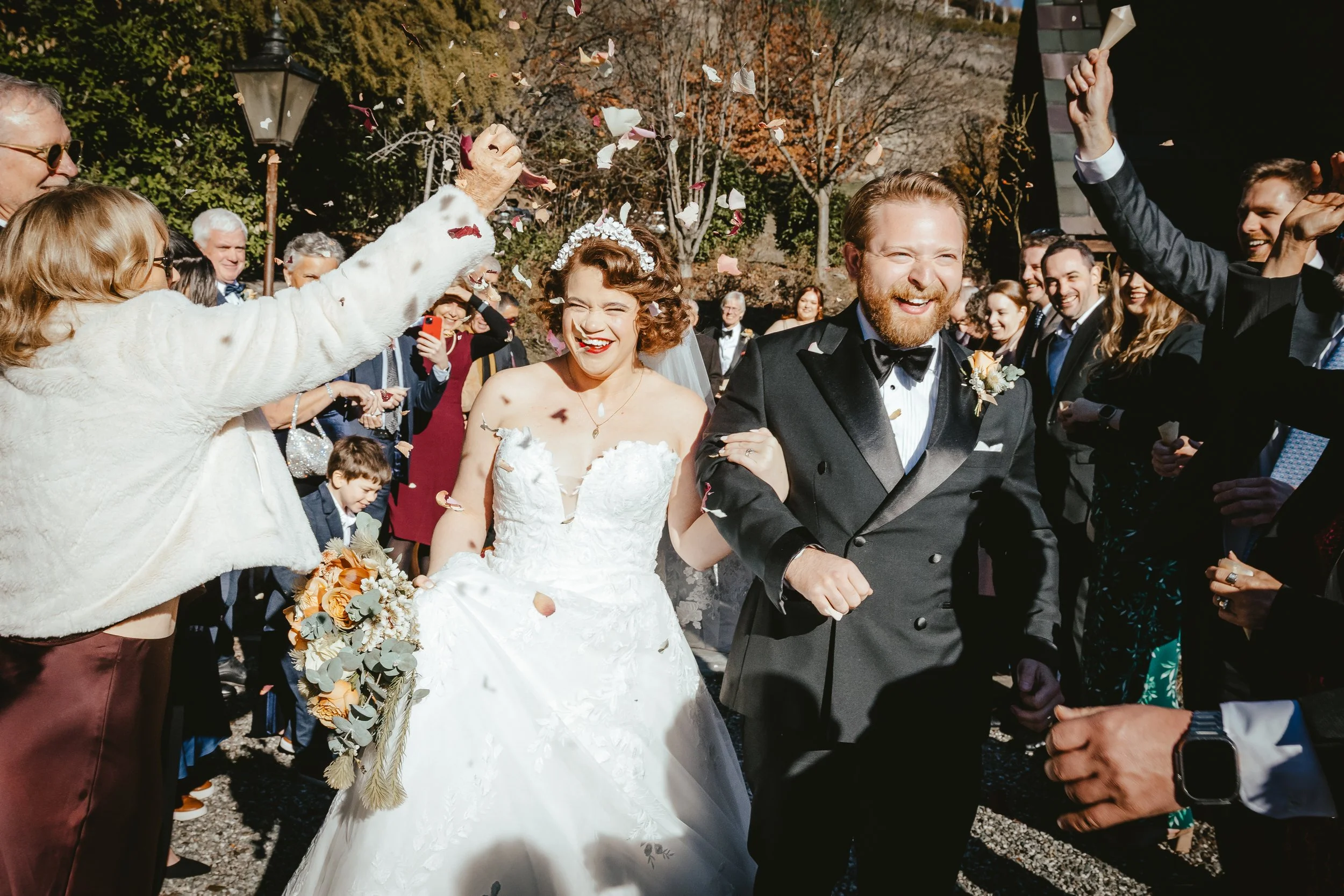 A bride and groom walking outdoors after their wedding, surrounded by smiling guests and confetti, during daytime with trees in autumn foliage.