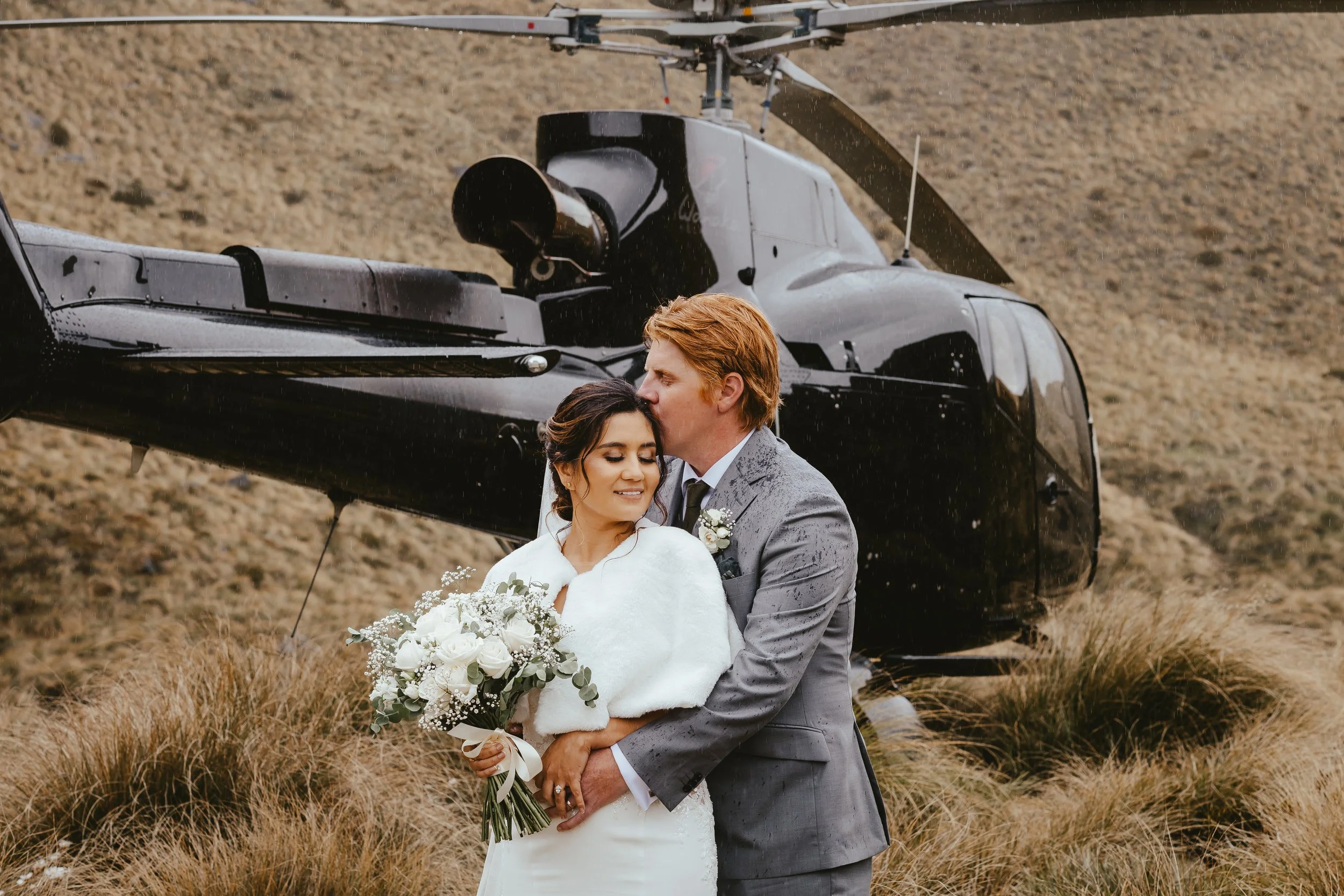 A newlywed couple in wedding attire standing outdoors in a field with tall grass. The groom is kissing the bride on the temple while holding a bouquet of flowers. A helicopter is in the background.