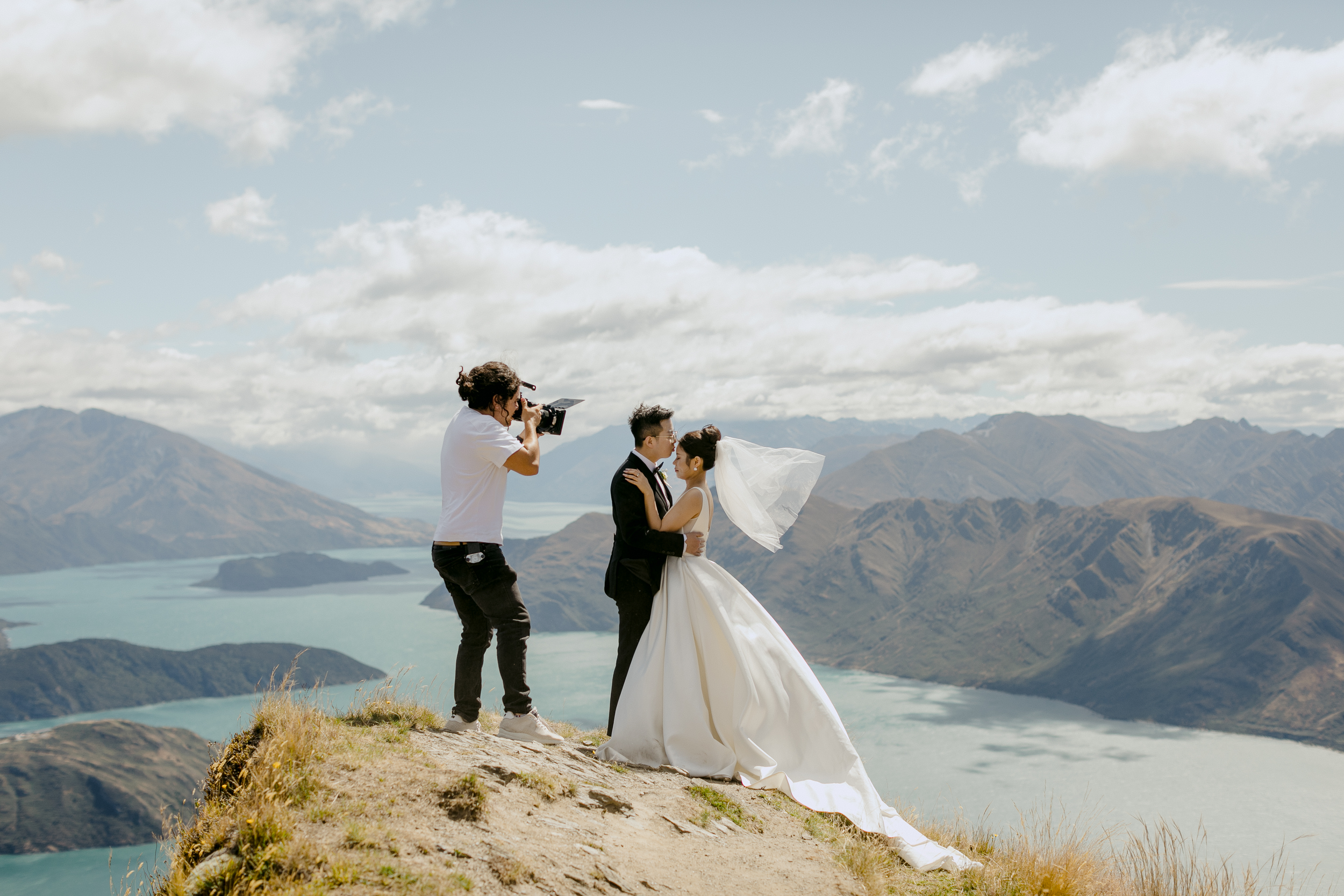 A bride and groom standing on a hilltop with mountains and a lake in the background, having their wedding photos taken by a photographer.