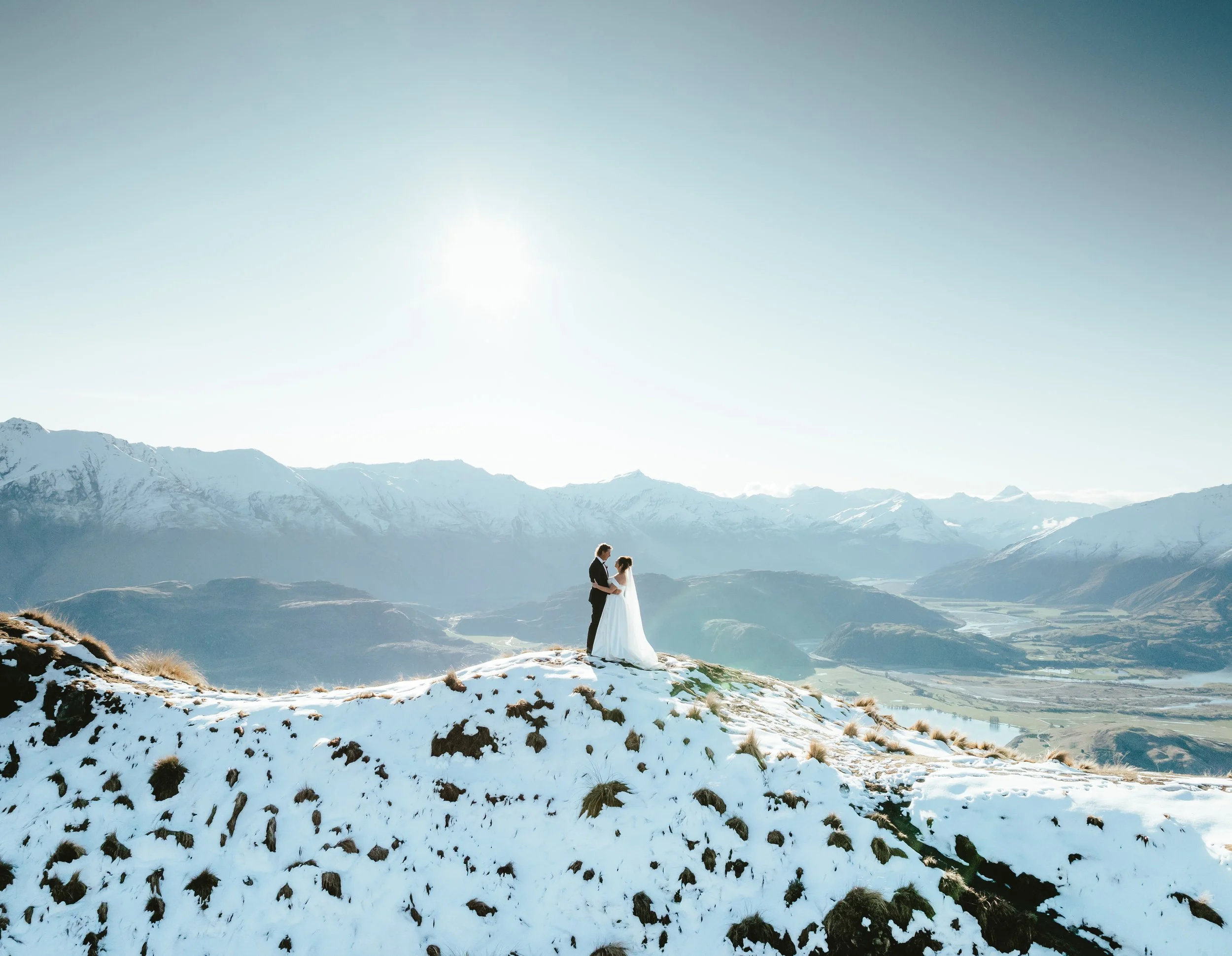 A couple in wedding attire standing on a snowy mountain ridge with snow-capped mountains in the background.