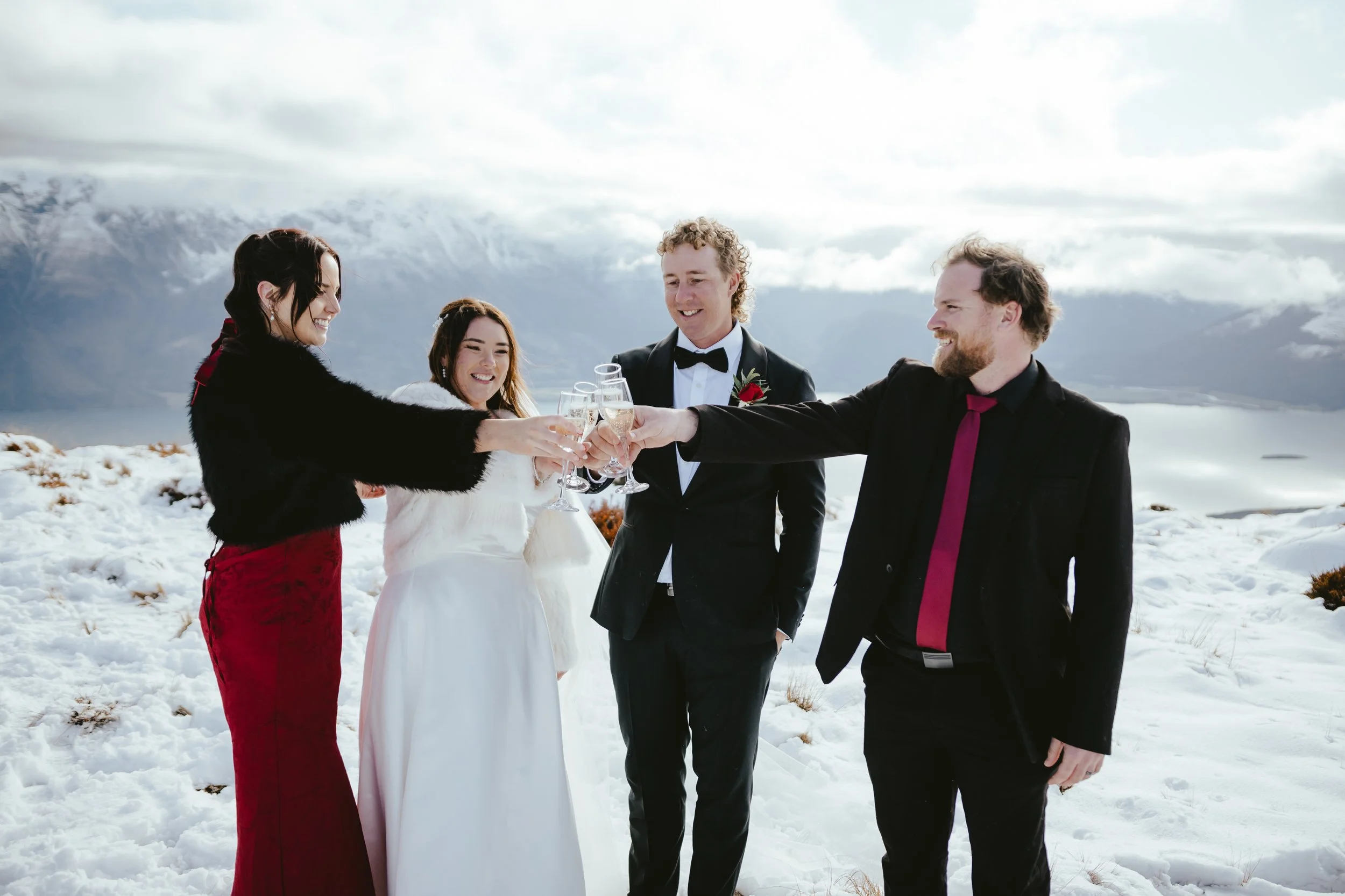 Group of five people in formal attire toasting with champagne glasses outdoors in a snowy landscape with mountains in the background.