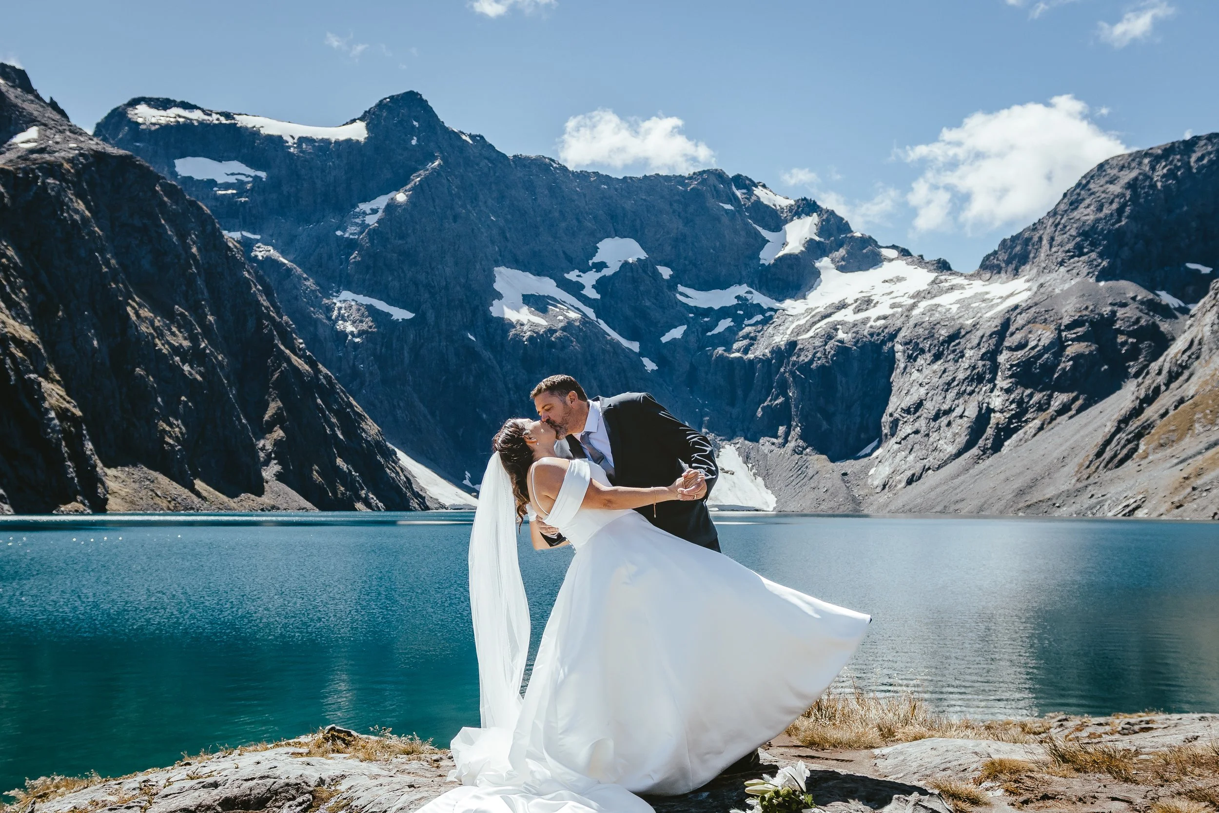 A bride and groom sharing a kiss by a mountain lake with snow-capped peaks in the background.