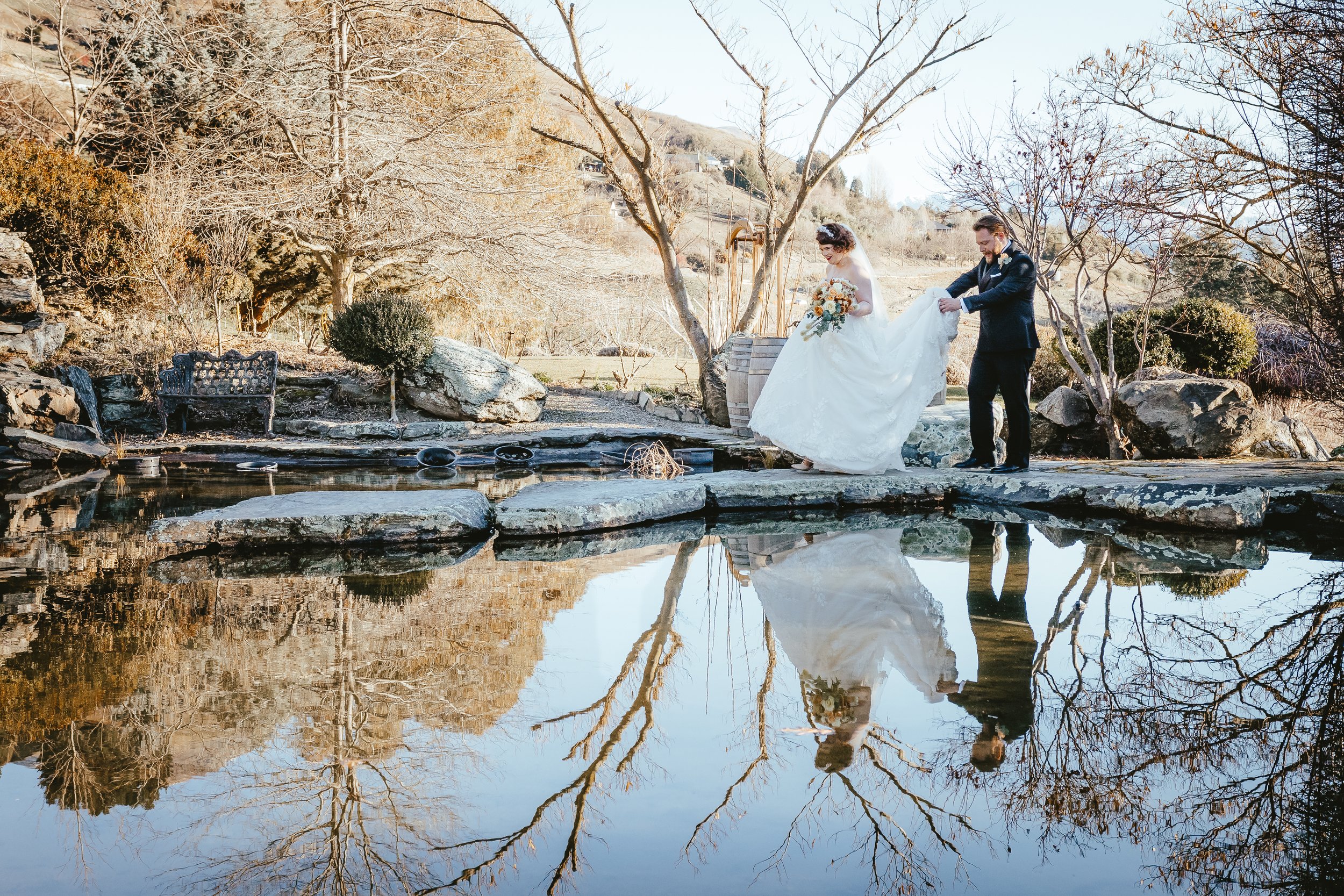 A couple dressed in wedding attire stands near a pond outdoors, holding hands and looking at each other. The bride holds a bouquet, and the groom is helping her with her dress. Trees with bare branches and rocks surround the water, reflecting in the 